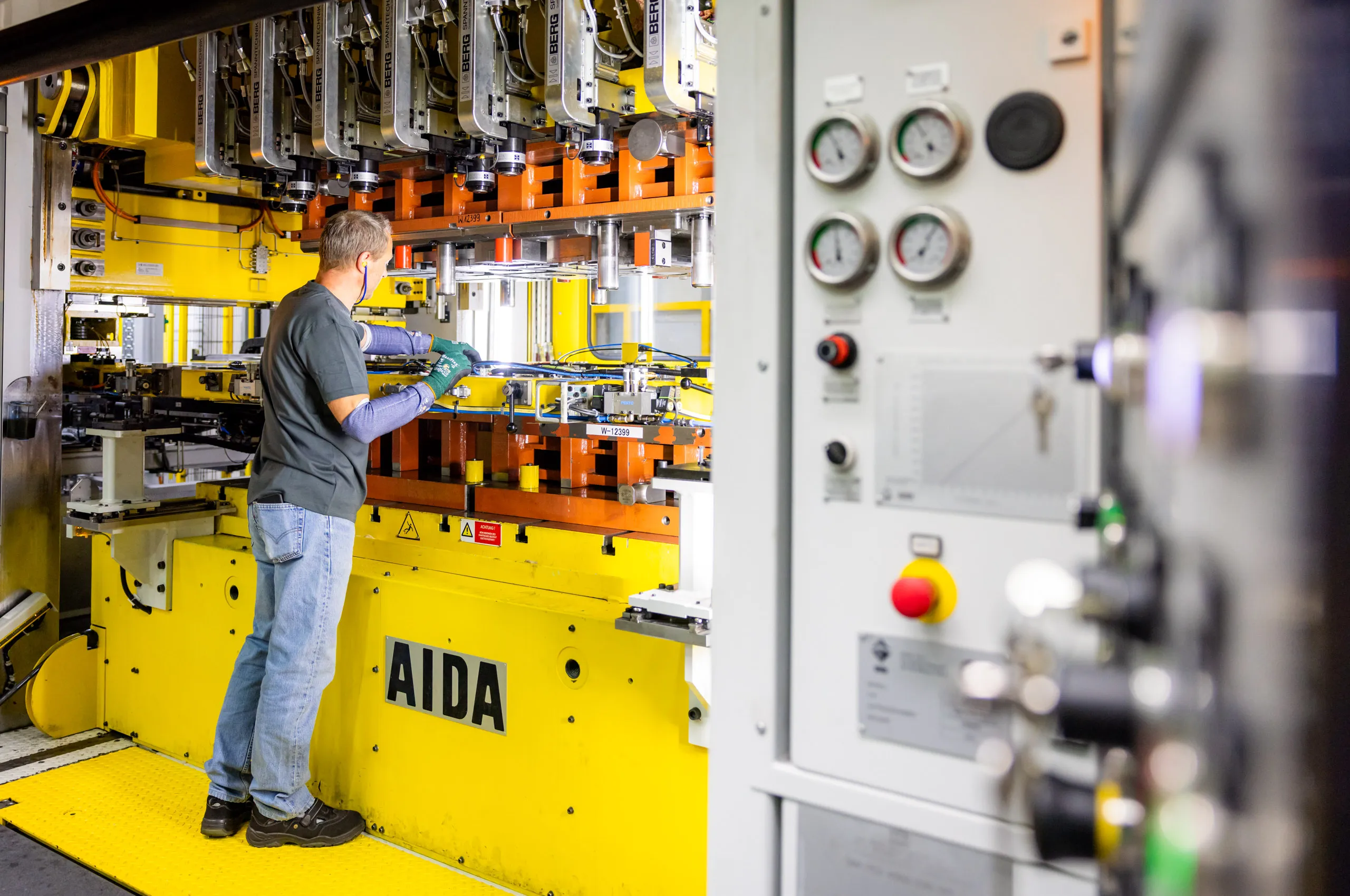 Ein Mann mit braunen kurzen Haaren steht seitlich gedreht an einer großen Presse und kontrolliert etwas an der Maschine. Die ganze Person ist in dem Foto zu sehen. Der Mitarbeiter trägt Sicherheitshandschuhe, Armschoner, eine blaue Jeans, ein graues T- Shirt und steht links im Bild auf einem gelben Untergrund. Auch die Maschine ist hauptsächlich gelb und orange. Rechts unscharf im Bild sind Schalter, Drehzahlmesser und Knöpfe der Maschine zu sehen