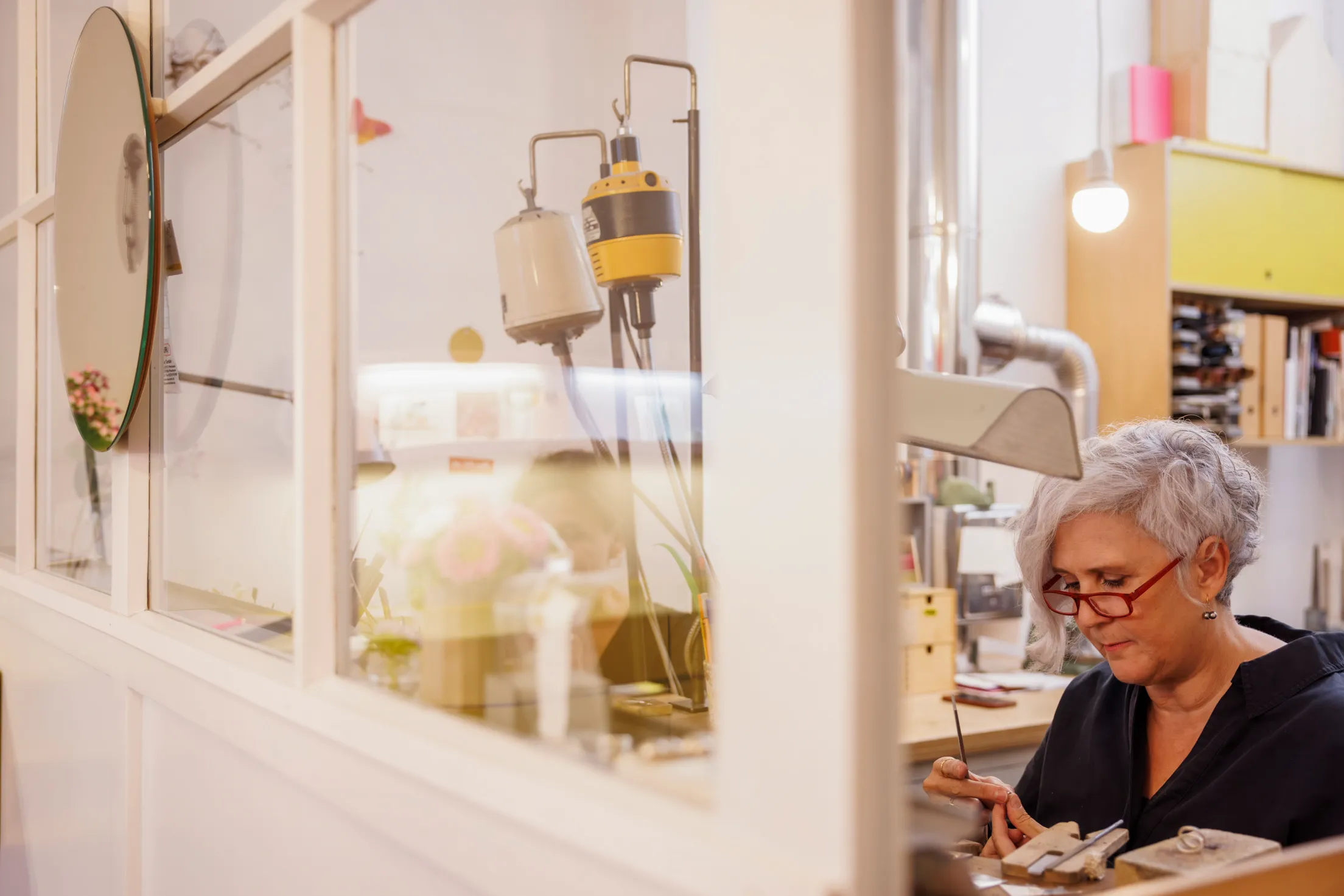 Eine weiße Werkstatttür mit Fenstern ist zu sehen, rechts davon sitzt eine Goldschmiedin mit hellgrauem Haar. Sie trägt ein schwarze Bluse und bearbeitet ein Schmuckstück