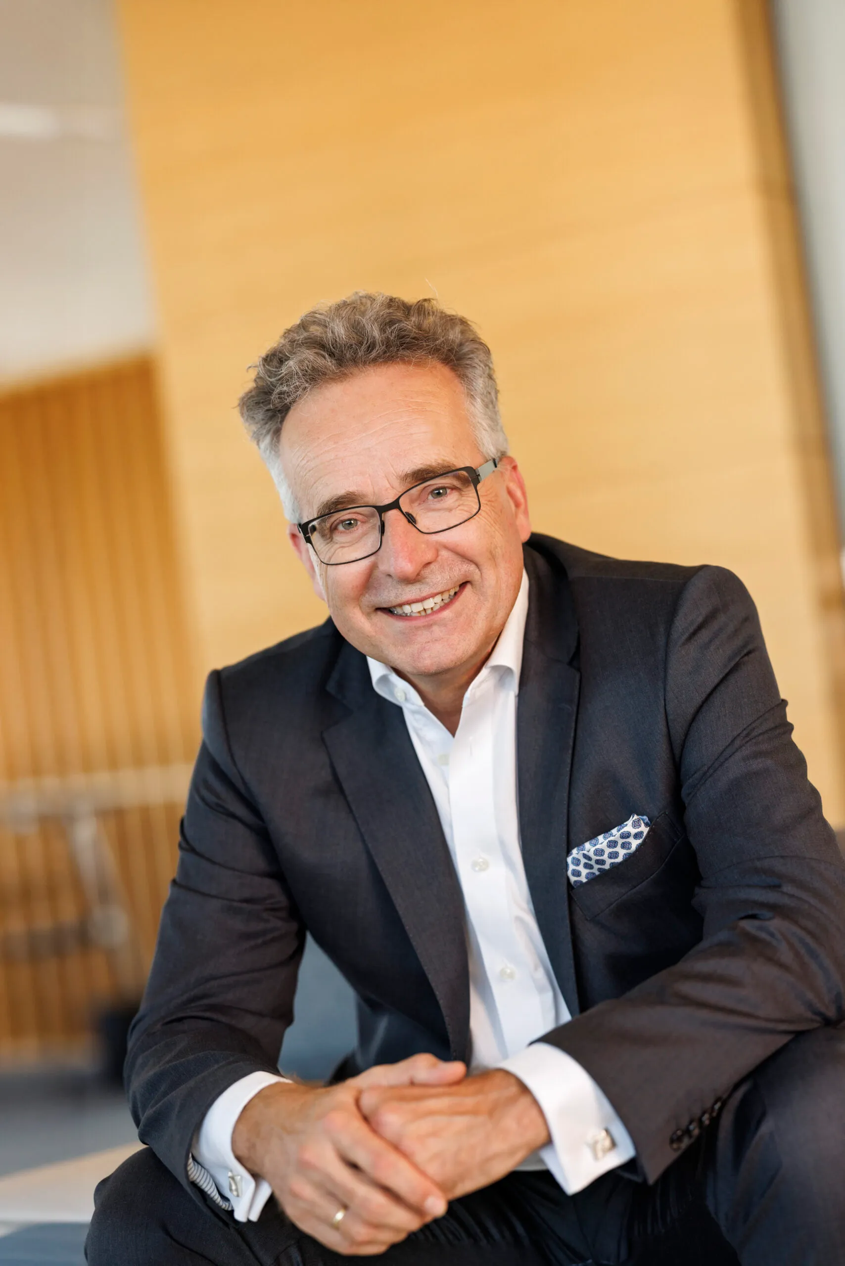 Ein Mann mit grauem, lockigen Haar und Brille ist im Sitzen zu sehen, er trägt einen dunkelgrauen Anzug, hat die Hände überschlagen und lächelt selbstbewusst in die Kamera