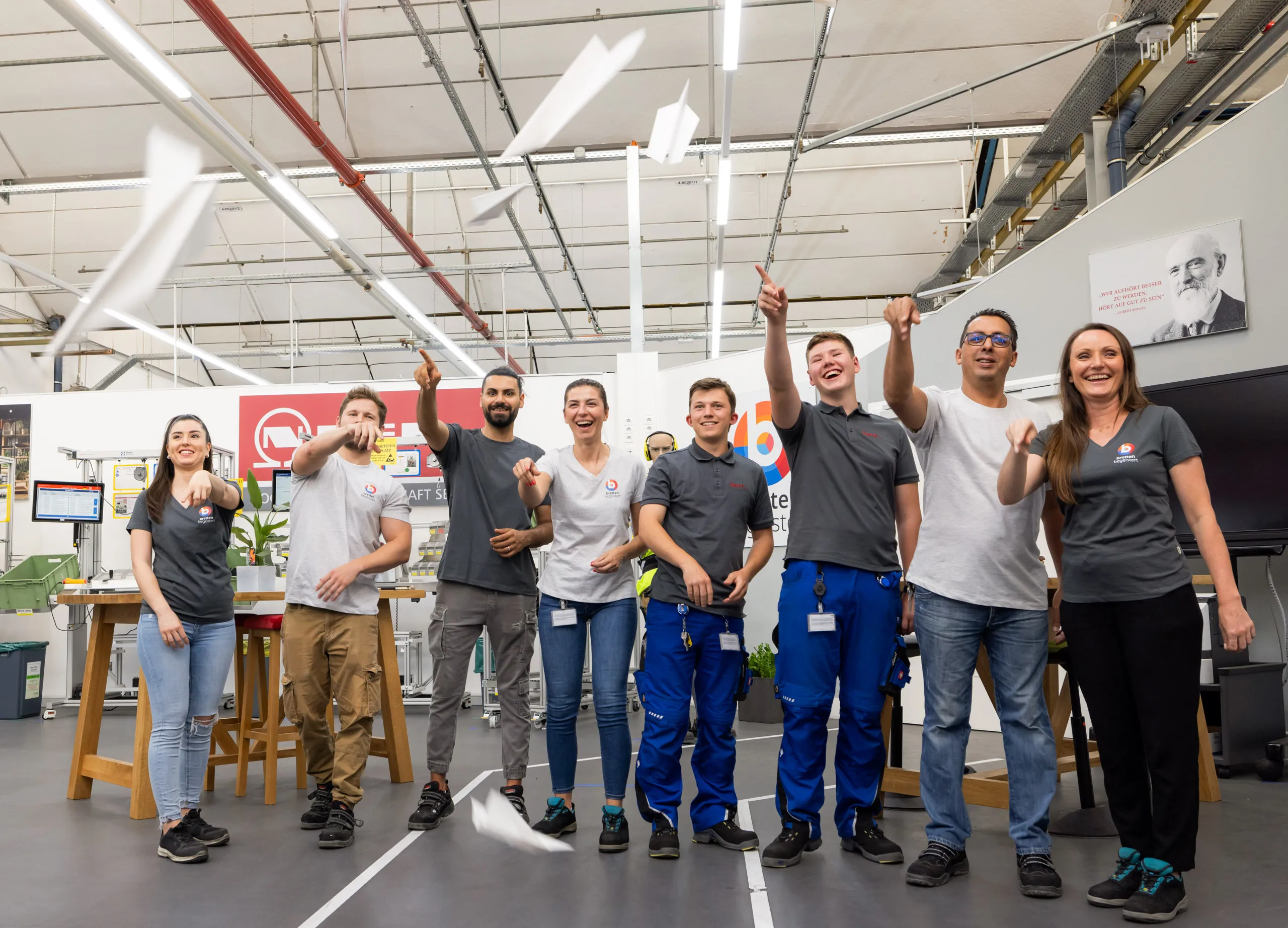 Eine Gruppe von acht jungen Menschen stehen in einer Reihe. Sie sind Auszubildende aus dem Werk in Bretten, tragen alle eine BSH Arbeitskleidung und haben weiße Papierflieger gebastelt. Diese lassen die jungen Menschen direkt in die Kamera fliegen und sie lachen dabei viel. Im Hintergrund ist die Trainingsakademie zu sehen, rechst davon ein Schwarz Weiß Foto von Robert Bosch.