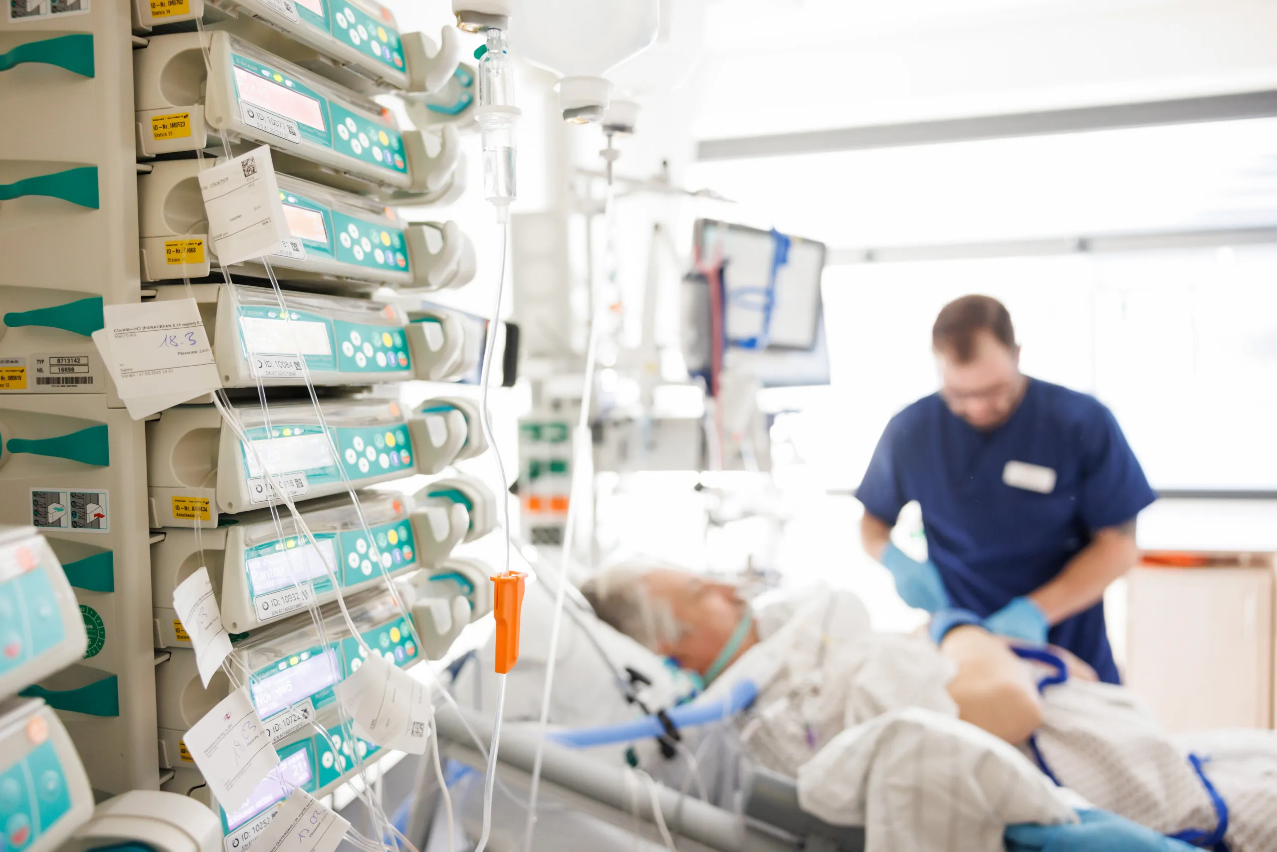 Das Foto zeigt ein sehr helles Zimmer auf der Intensivstation. Im linken Drittel sind im Anschnitt scharf die Überwachungsmonitore mit türkisfarbenen Knöpfen zu sehen mit verschiedenen Displays, zusätzlich noch Infusionen und Schläuche. Zwei Drittel des Bildes zeigen in der Unschärfe ein Patientenbett mit einem grauhaarigen, älteren Herrn und Schläuchen. Bei ihm steht ein Pfleger in einem blauen Kasack und kontrolliert etwas.