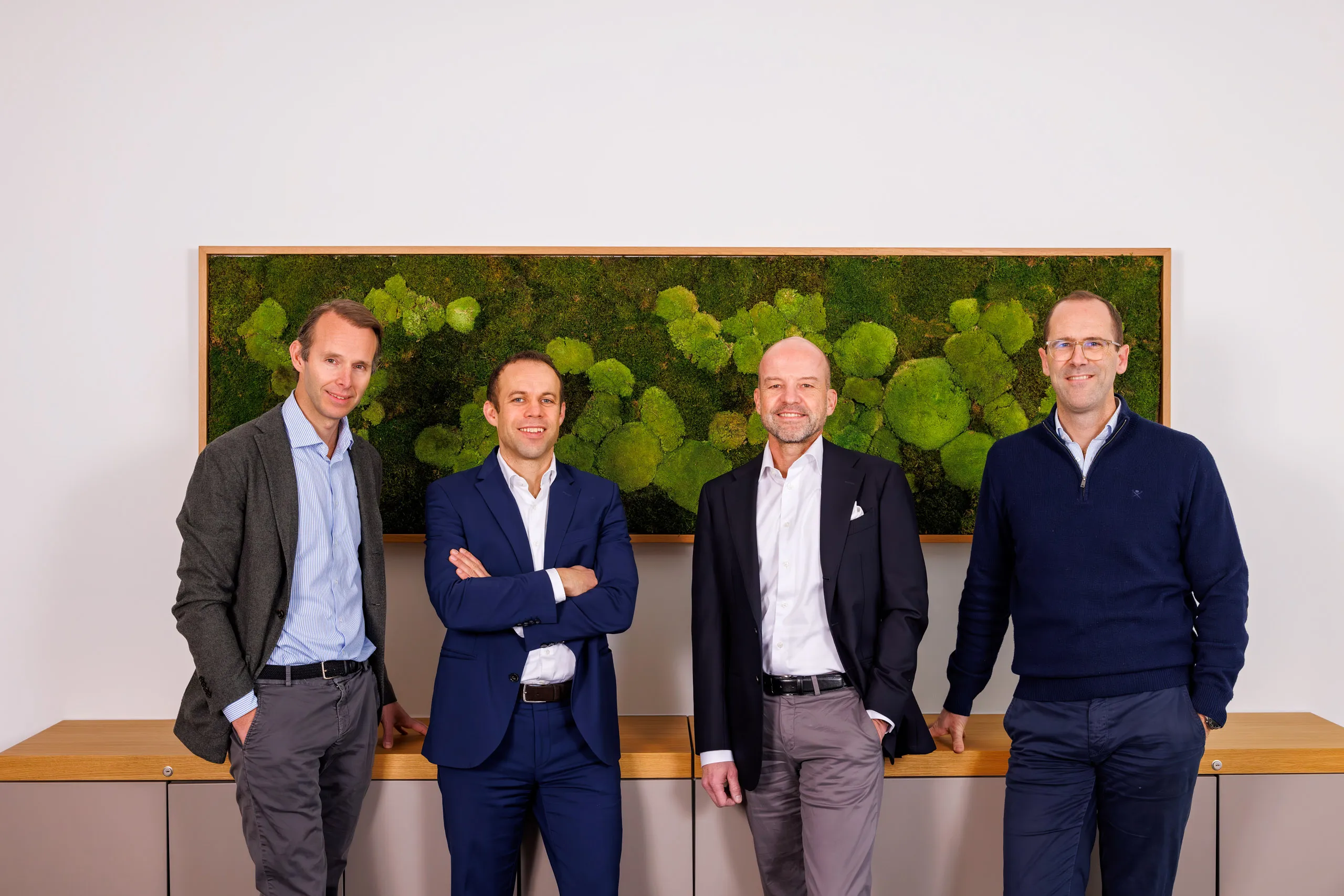 Vier Geschäftsführer der Firma stehen nebeneinander an einem Sideboard mit Holzplatte gelehnt. Sie tragen alle ein Businessoutfit und lächeln in die Kamera. Im Hintergrund der Herren ein grünes Bild mit Moos in einem Holzrahmen und einer weißen Wand.