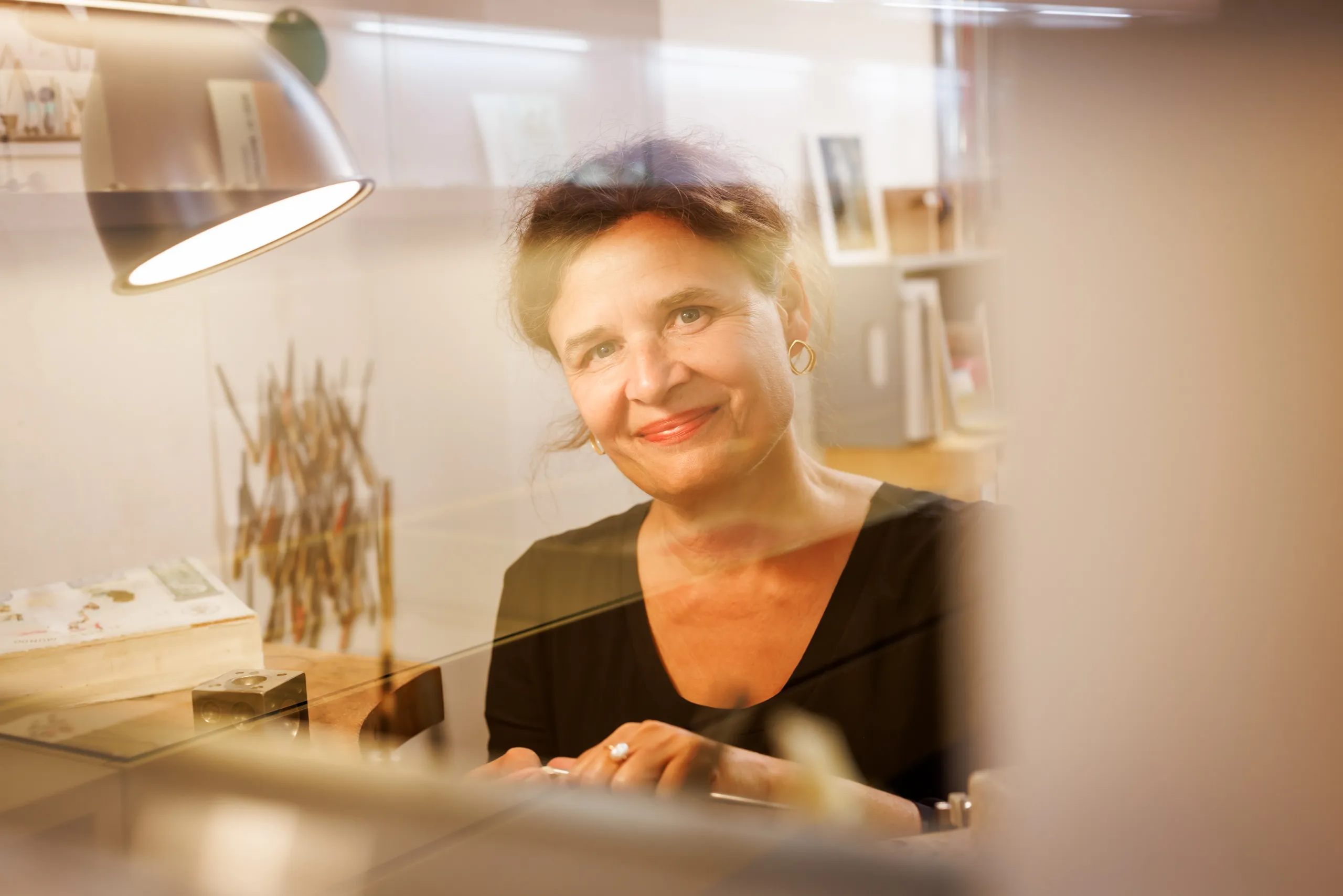 Eine Goldschmiedin bei der Arbeit in ihrer Werkstatt. Blick in die Kamera. Foto wurde durch eine Glasscheibe aufgenommen. Die Dame trägt einen Zopf, hat braune Haare und und Goldohrringe. Sie hat ein schwarzes T-Shirt an und lächelt in die Kamera.