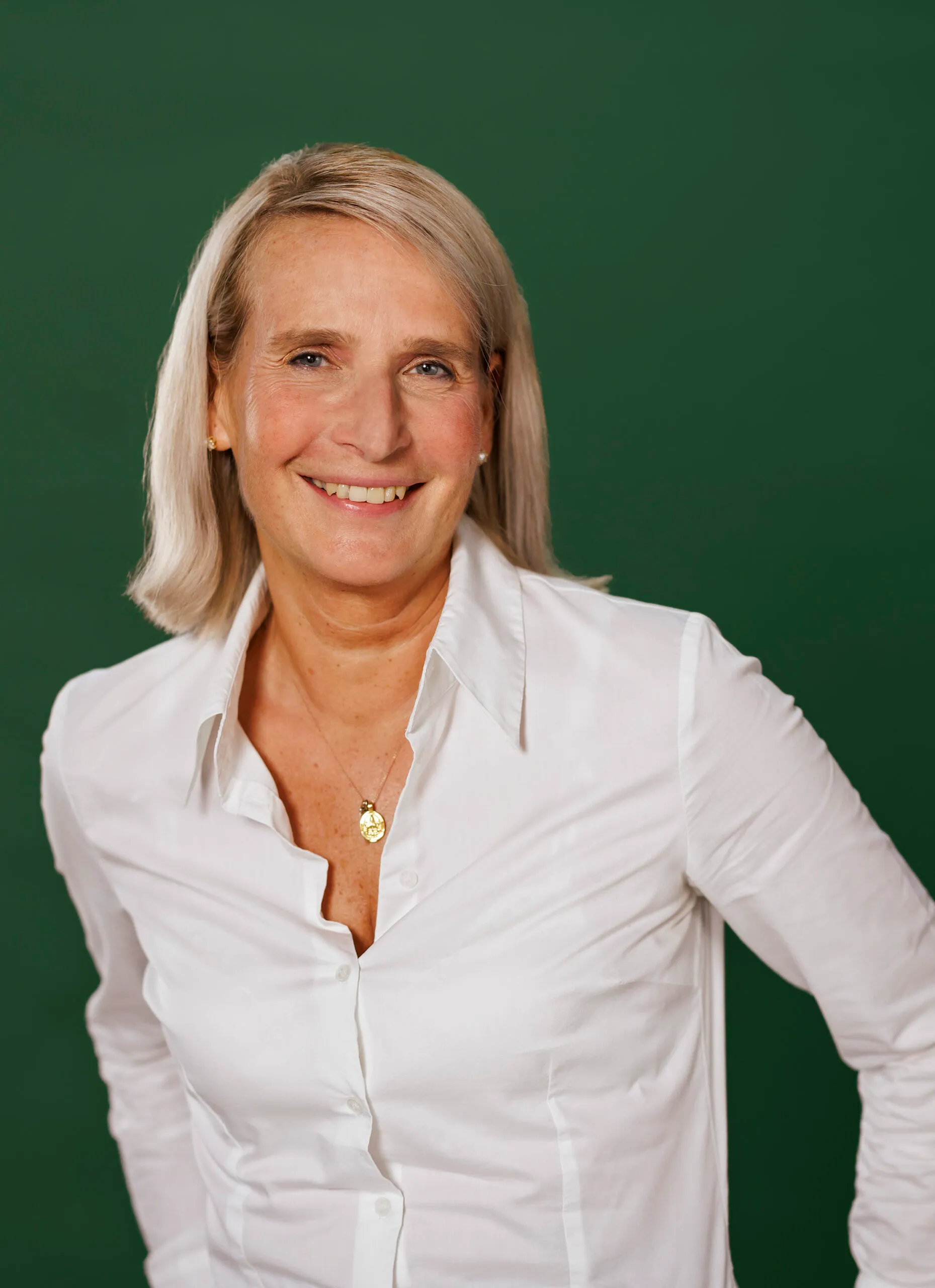Frau mit kinnlangem hellen Haar lächelt selbstbewusst in die Kamera. Der Hintergrund ist dunkelgrün und sie trägt eine weiße Bluse.