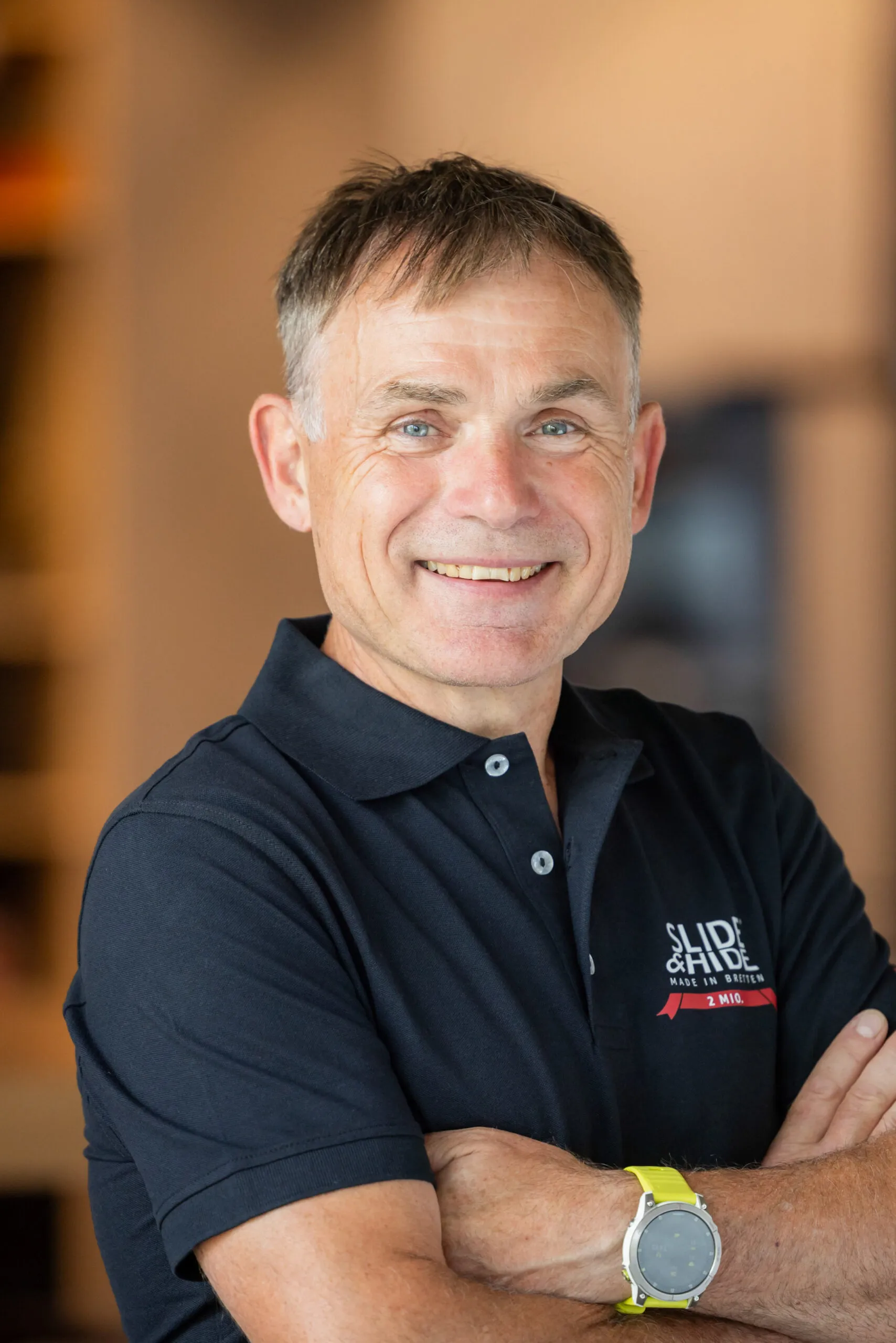 Portraits von einem Mann mittleren Alters mit verschränkten Armen und einem blauen Polohemd. Er trägt eine gelbe Armbanduhr und lacht in die Kamera. Er hat braune und leicht ergraute, kurze Haare und steht vor einem sehr unscharfen, orangefarbenen Hintergrund.