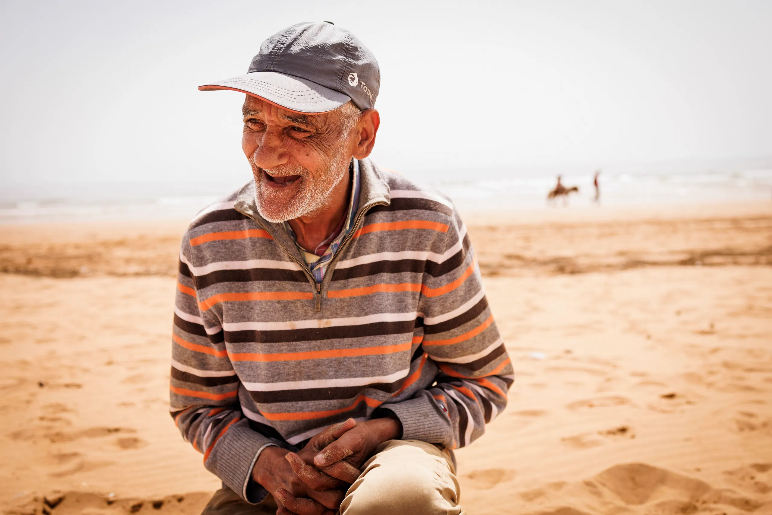Ein älterer marokkanischer Mann ohne Zähne, mit gestreiftem Pullover und einem grauen Cap sitzt in der Hocke am Strand und lacht zur Seite. Im Hintergrund ist unscharf das Meer mit einem Reiter zu sehen.