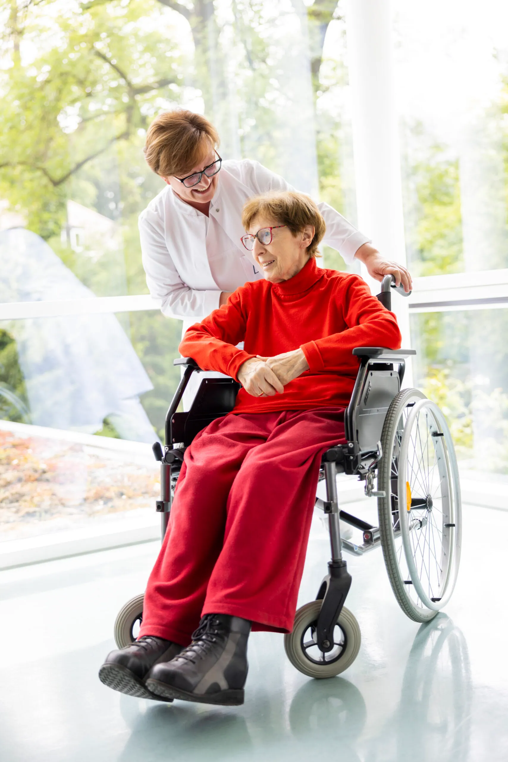 Eine Ärztin schiebt einen Rollstuhl uns beugt sich sehr fürsorglich zu der Patientin im Rollstuhl. Diese trägt einen knalligen roten Rollkragenpullover und eine rote Hose.