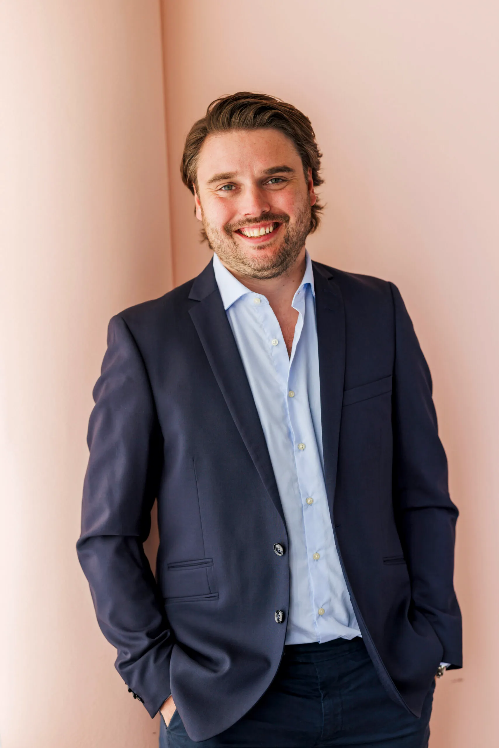 Ein jüngerer Mann schaut lächelnd in die Kamera. Er steht vor einer rosafarbenen Wand und trägt ein blaues Jacket mit einem hellblauen Hemd.