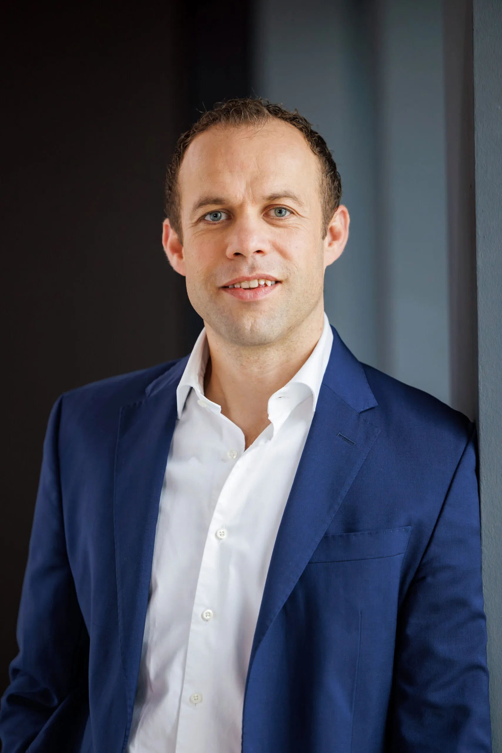 Portraits von der Geschäftsführung, ein Mann mittleren Alters mit braunem kurzem Haar. Er trägt ein weißes Hemd, ein blaues Sakko und schaut in die Kamera. Im Hintergrund dunkle, unscharfe Strukturen in Schwarz und Grautönen.