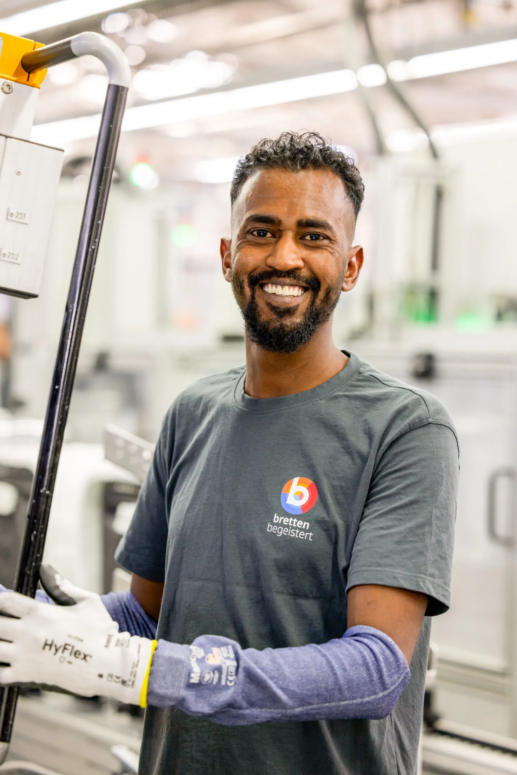 Ein farbiger Mitarbeiter mit schwarzem Haar und Bart, trägt ein dunkelgraues BSH Werksshirt, weiße Sicherheitshandschuhe und blaue Armschoner. Er lacht in die Kamera und ist mit dem Oberkörper zu sehen, leicht nach links gedreht. Er ist beim Arbeiten und hält eine schwarze Stange fest. Der Hintergrund ist sehr hell und unscharf in einer Fabrikhalle.