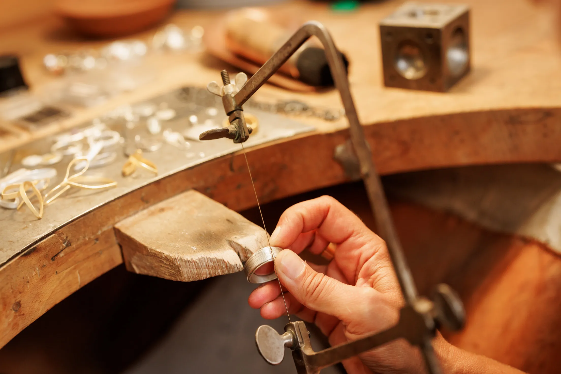Blick von oben auf eine Säge und eine Hand, die einen silbernen Ring hält, der gerade bearbeitet wird. Sehr warmes Licht auf einer Holzplatte mit weiterem Schmuck Material in der Unschärfe.