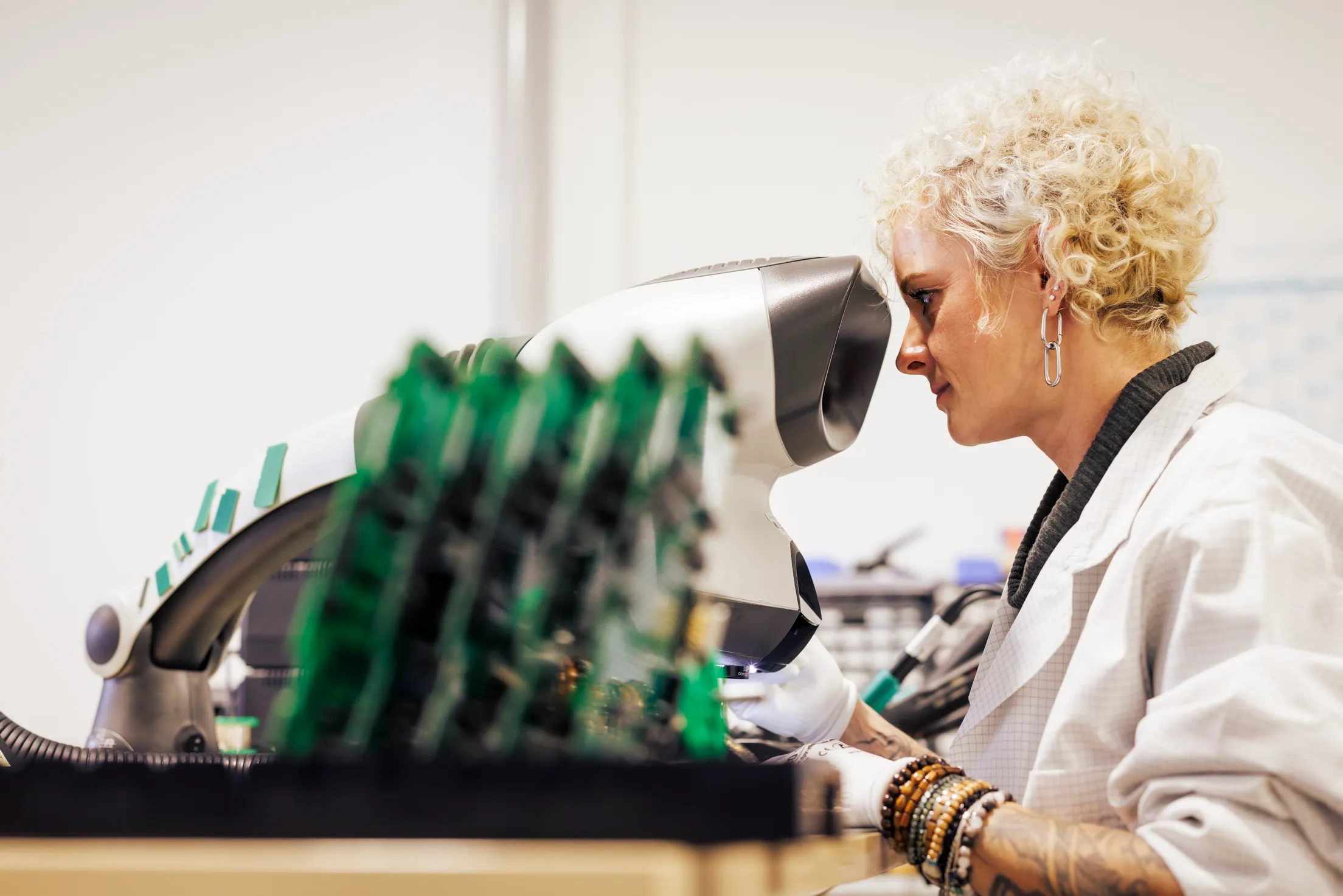 Eine blond gelockte Frau ist von der Seite zu sehen, nur ihr Oberkörper und sie schaut durch ein Mikroskop. Sie trägt einen weißen Kittel und Handschuhe