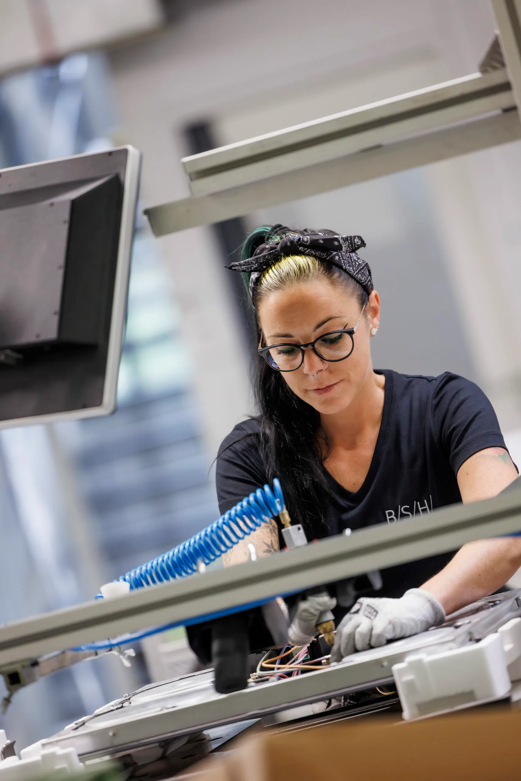 Eine Mitarbeiterin steht am Fließband, sie ist schwarz gekleidet mit einem schwarzen Kopftuch und schraubt an einer Herdplatte. Sie wurde durch ein Gerüst durch fotografiert.