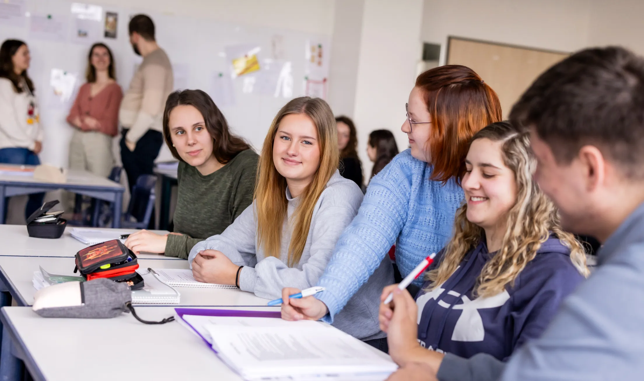Eine Reihe von Schülerinnen ist in einem Klassenzimmer fotografiert. Sie sitzen in einer Reihe an hellen Schulbänken und eine Schülerin schaut direkt in die Kamera, die anderen schauen sich an. Sie trägt ein hellgraues Sweatshirt und hat mittelblonde, glatte, lange Haare. Auf dem Tisch liegen Hefte, Blöcke und Federmäppchen. Links hinten im Bild stehen drei Schüler und Schülerinnen zusammen und unterhalten sich.