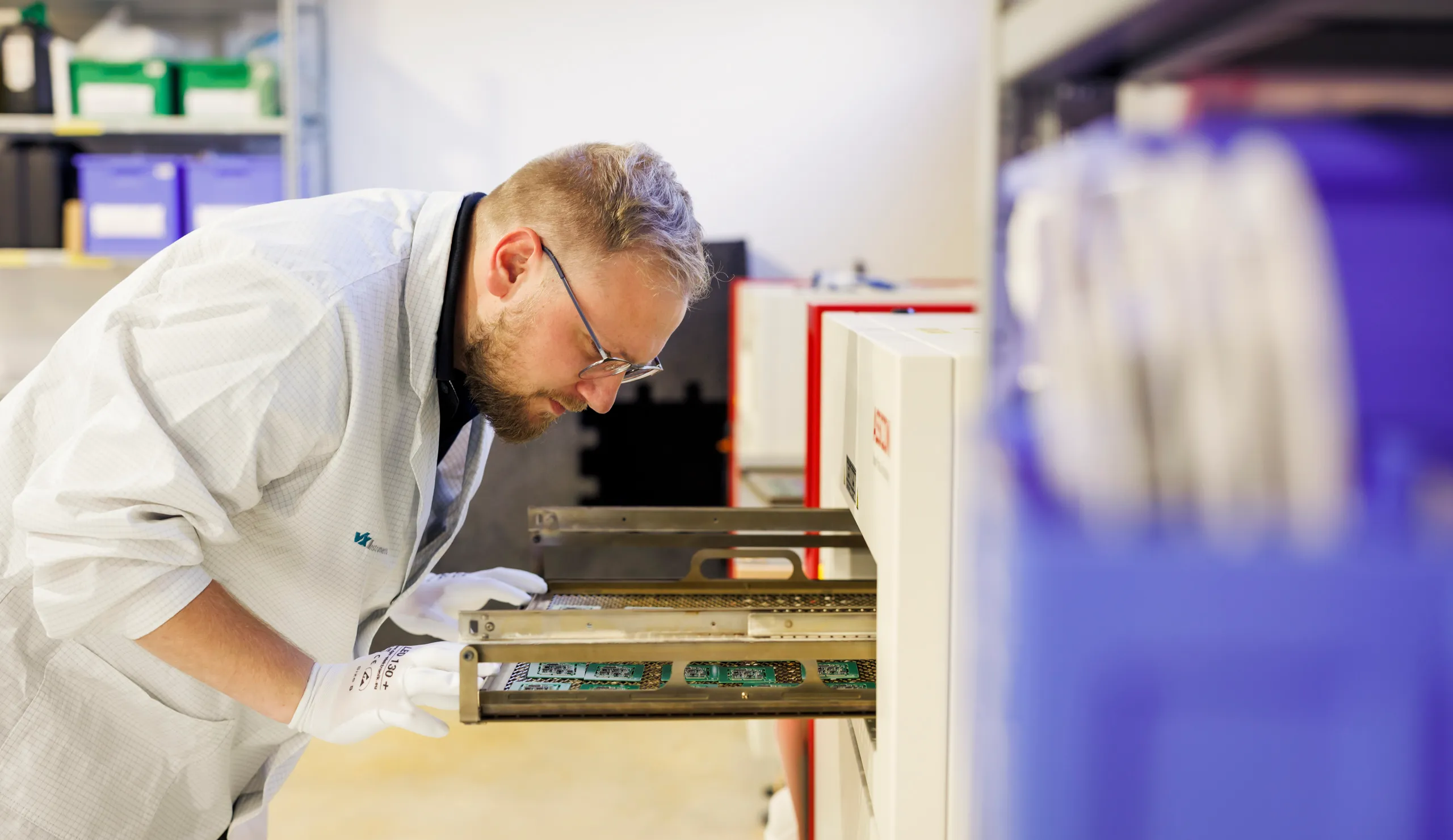 Ein Arbeiter mit weißem Kittel und Brille ist von der Seite zu sehen, nur sein Oberkörper ist gezeigt. Er zieht gerade eine Schublade aus einer Maschine und er prüft den Inhalt.