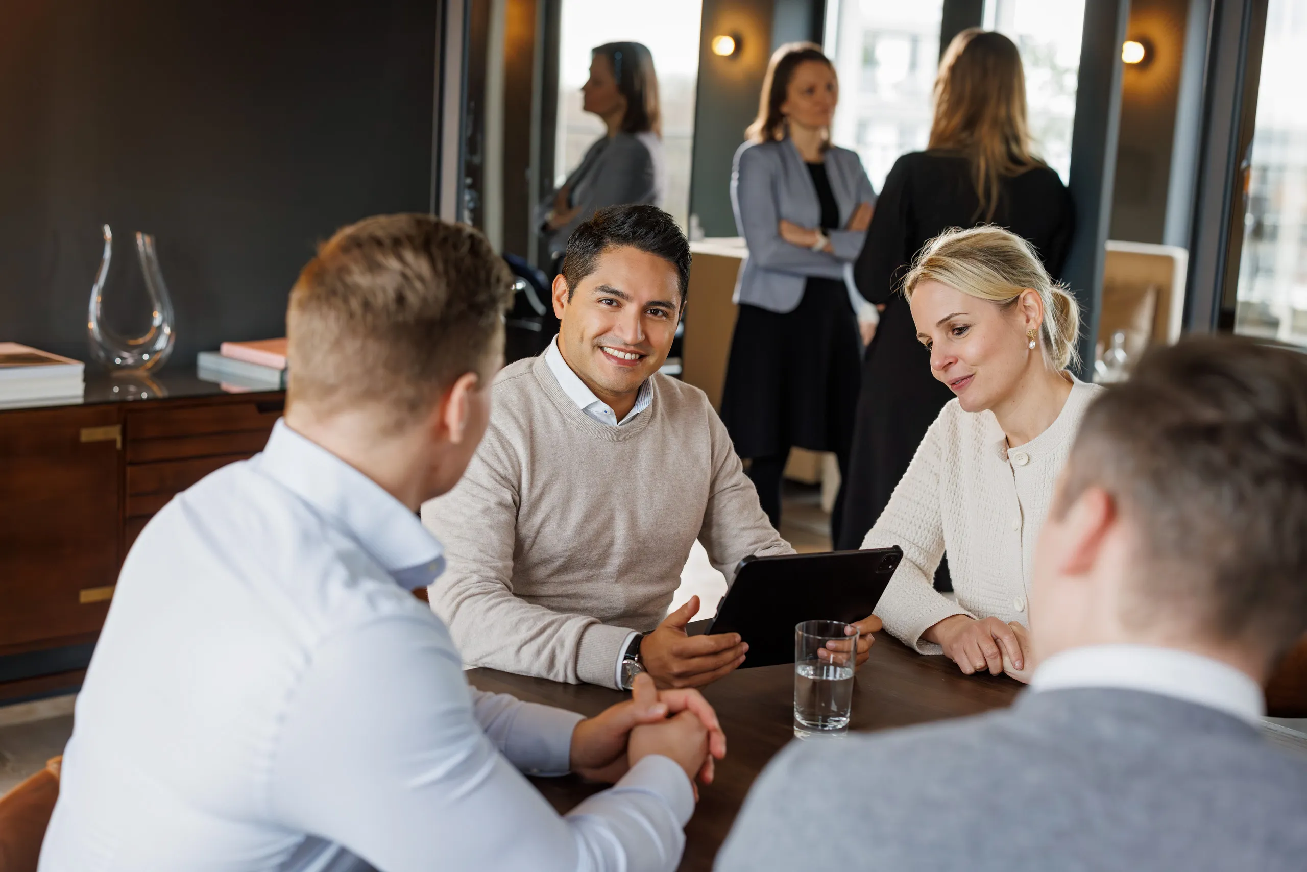 Eine Gruppe von vier Personen sitzen an einem Konferenztisch mit einem Tablet und einem Glas Wasser auf dem braunen Tisch. Sie besprechen etwas. Zwei Personen sitzen mit dem Rücken zu der Kamera in der Unschärfe, sie trage beide helle Kleidung. Fokus ist auf zwei Personen, die zu der Kamera gewandt sind. Mittig ein Mann mit hellem Hemd und beigen Pullover hält das Tablet. Links von ihm sitzt eine Dame mit blondem Pferdeschwanz, hellbeige Jacke und sie schaut auf den Bildschirm. Im Hintergrund sind unscharf zwei Mitarbeiterinnen im Businessoutfit im Gespräch, links im Bild ein Sideboard aus Holz.
