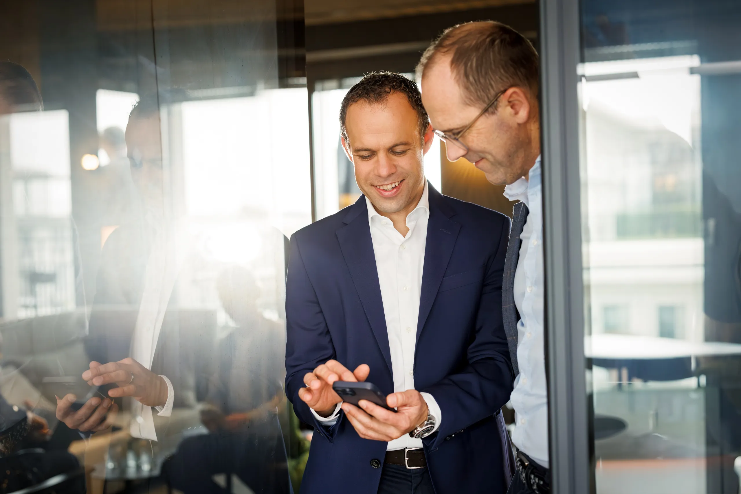 Zwei Männer im Business Outfit stehen zwischen zwei Glastüren und schauen beide lächelnd in ein Mobiltelefon. Der linke Mann trägt ein dunkelblaues Jacket, weisses Hemd und hat braune kurze Haare. Der andere im Anschnitt mit Brille, braunem, kurzem Haar, hellblauem Hemd und Jacket. Bei sind mittleren Alters. Viel Spiegelung im Glas und Gegenlicht von hinten durch die Glasscheiben.