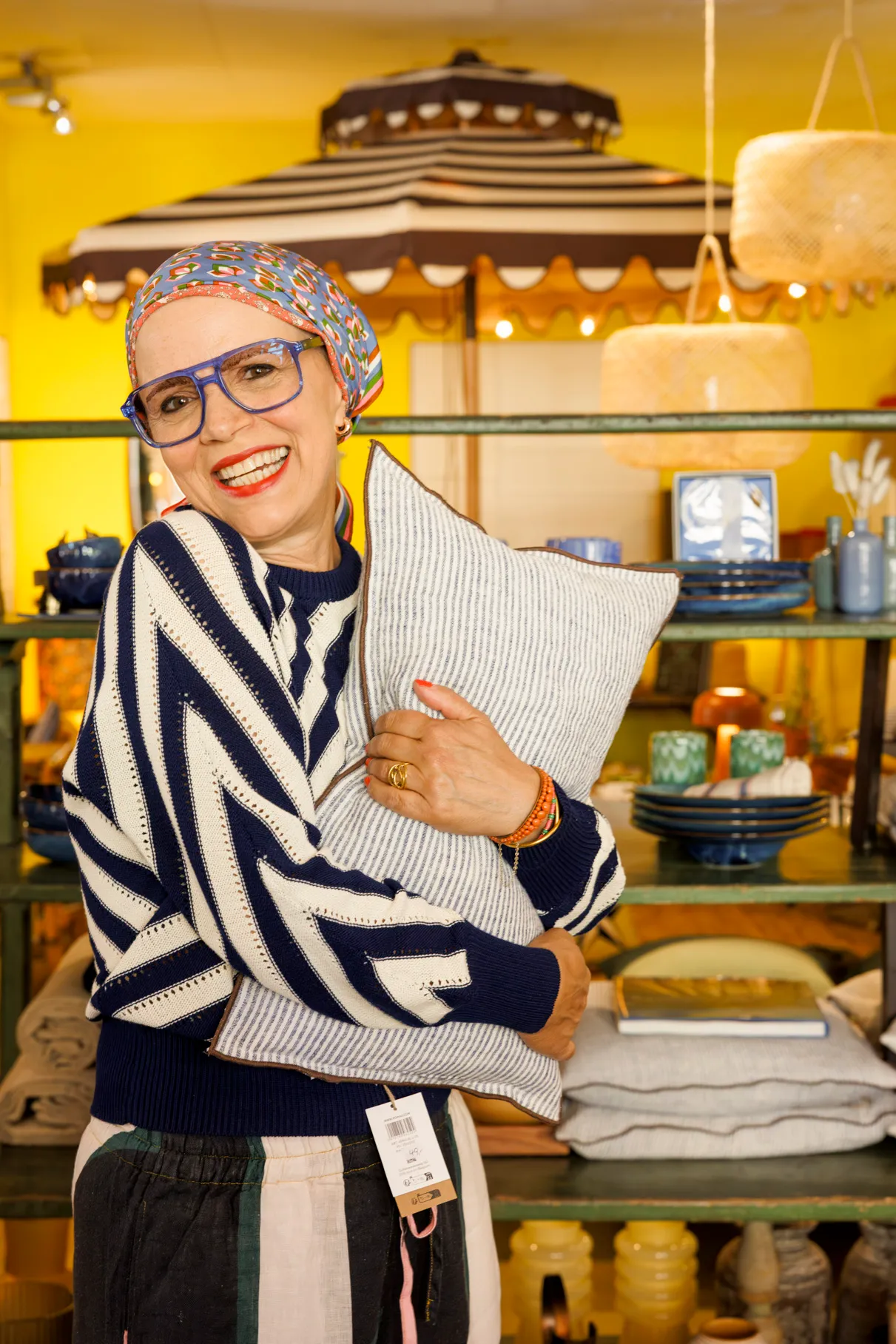 Eine Frau mit Kopftuch und auffälliger, blauer Brille und gestreiftem Pullover steht in einem Laden mit Wohnaccessoires. Sie lachst sehr symphatisch in die Kamera