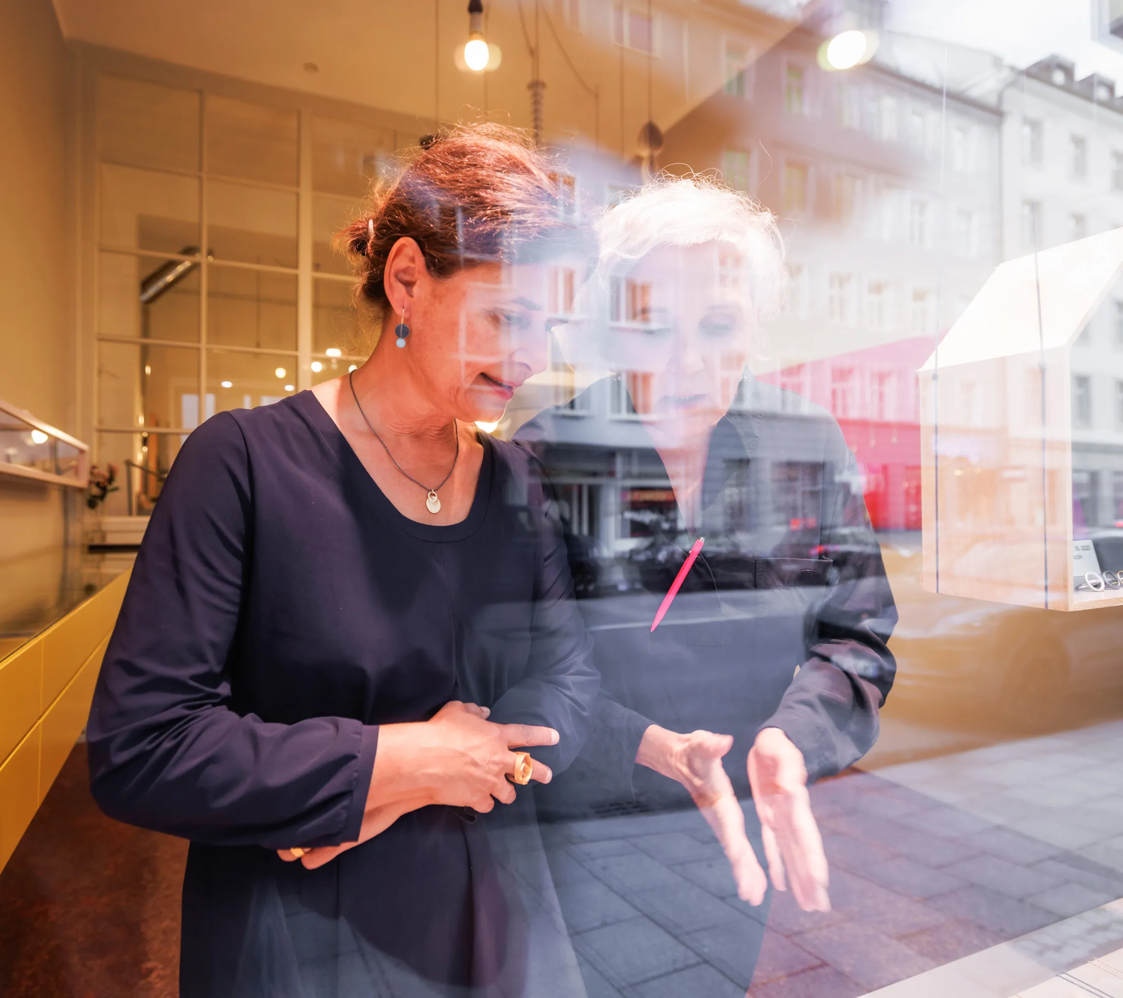Zwei Damen in dunklen Kleidern sind durch das Schaufenster fotografiert und durch die Spieglung im Fenster nicht klar zu erkennen. Sie sprechen und betrachten den Schmuck dabei, der in dem Schaufenster ausgestellt ist. Der Schmuck ist aber nicht zu sehen. Die Dame links hat braunes, die Andere hellgraues Haar. Im Hintergrund unscharf der Schmuckladen erkennbar mit viel Licht.