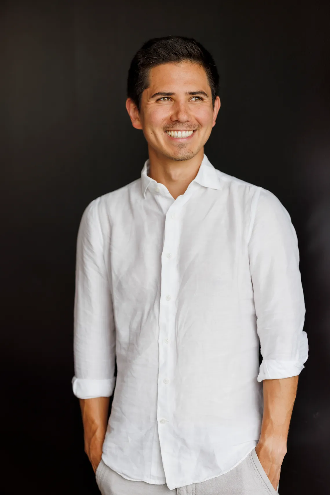 Ein junger Mann mit schwarzem Haar und weißem Hemd steht vor einem schwarzen Hintergrund. Er schaut lächelnd zu seiner linken Seite aus dem Bild.