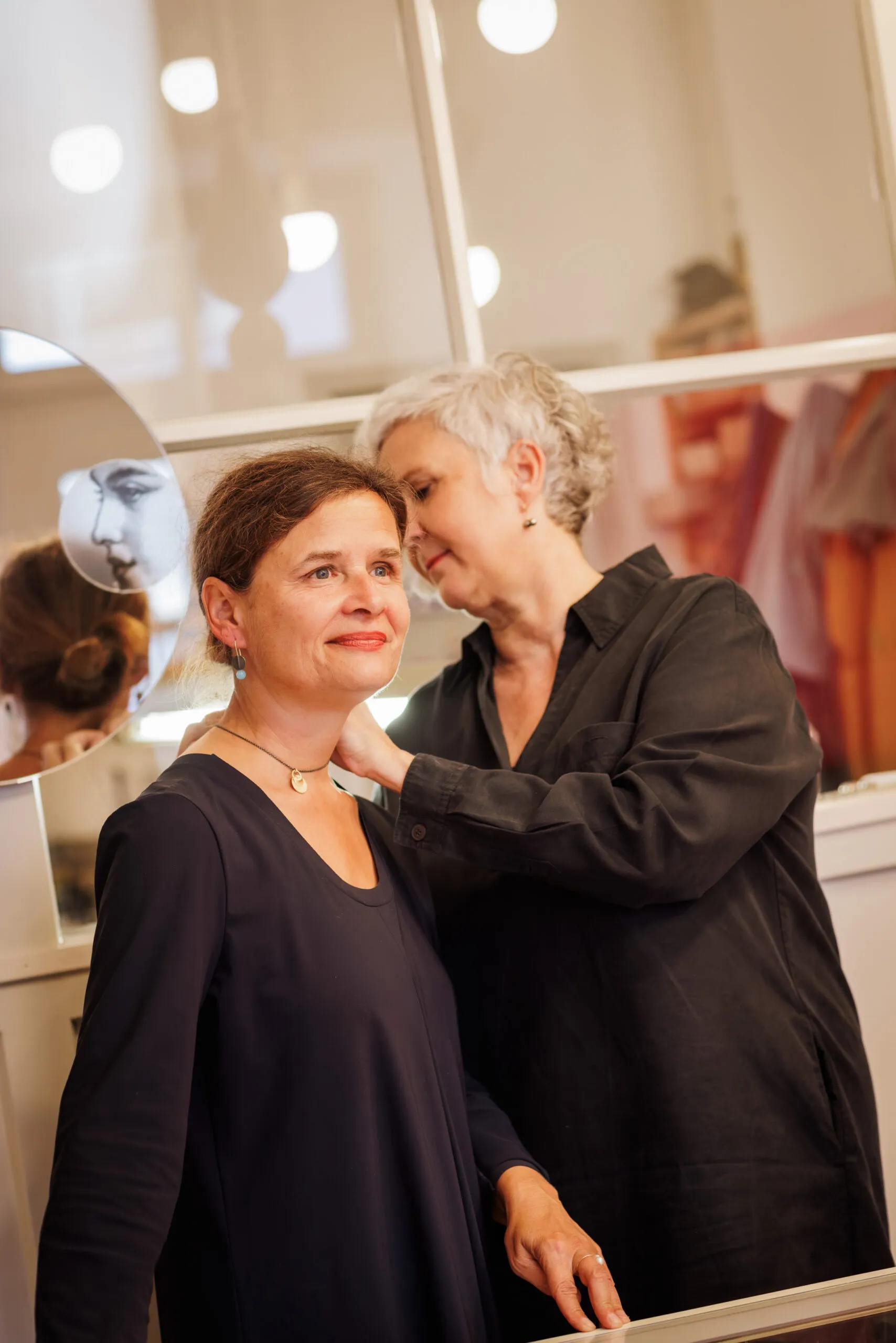 Zwei Goldschmiedinnen stehen zusammen vor einem Fenster im Schmuckladen, links ein Spiegel im Anschnitt. Die eine bindet gerade der anderen Dame eine Halskette um den Hals. Beide tragen dunkle Kleider und schauen nicht in die Kamera. Die Frau links hat braune Haare in einem Zopf, die andere Dame kurzes, graues Haar.