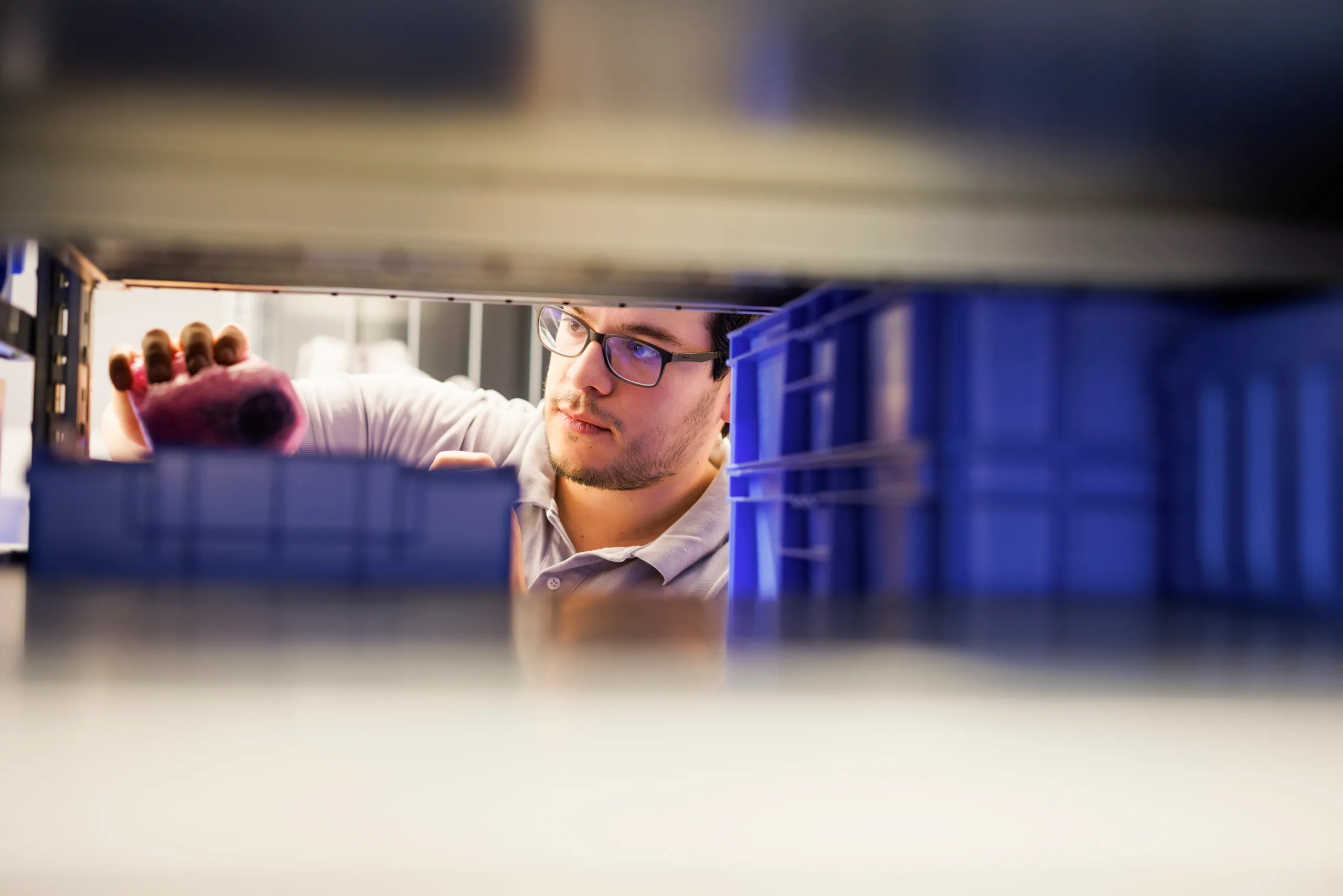 Ein Mann mit Brille holt etwas aus einer blauen Kiste. Er ist aus einer interessanten Perspektive durch blaue Kiste in einem Regal fotografiert.