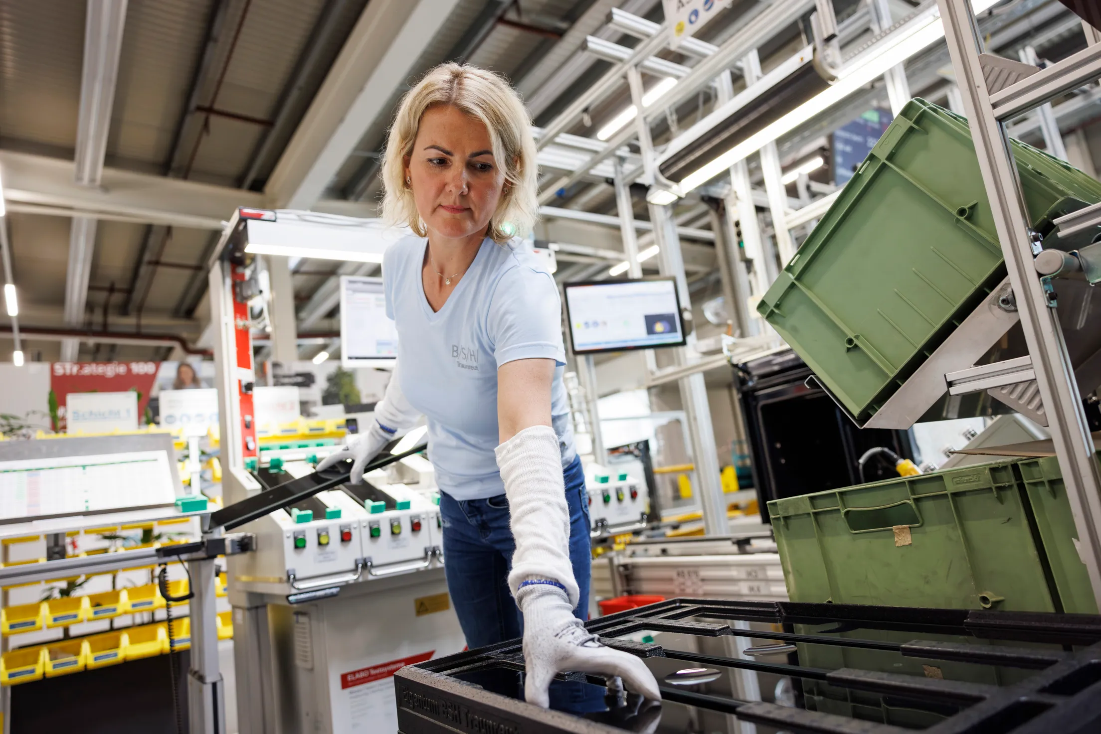 Eine Mitarbeiterin mit hellblauen Shirt, hat blonde kinnlange Harre und nimmt eine Blende von einem Backofen in die Hand. Sie trägt weiße Handschuhe und das Foto ist sehr weit von unten aufgenommen.