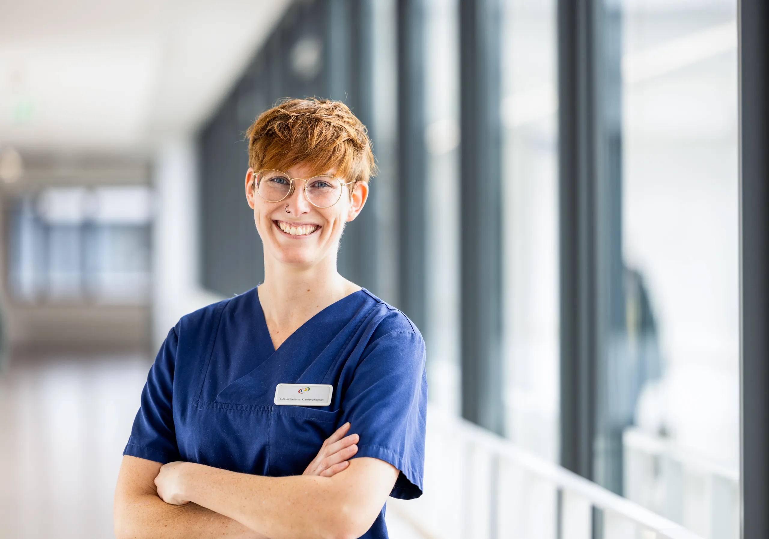 Eine jüngere, sehr sympathische Krankenschwester trägt einen dunkelblauen Kasack und hat ihre Arme verschränkt. Sie schaut mit einem intensiven Lachen in die Kamera, hat kupferrotes, kurzes Haar, eine auffällige goldene Brille und im rechten Nasenloch einen Ring. Sie steht in einem hellen Klinikgang mit dunkelgrauen, großen Fenstern, welcher unscharf fotografiert ist.