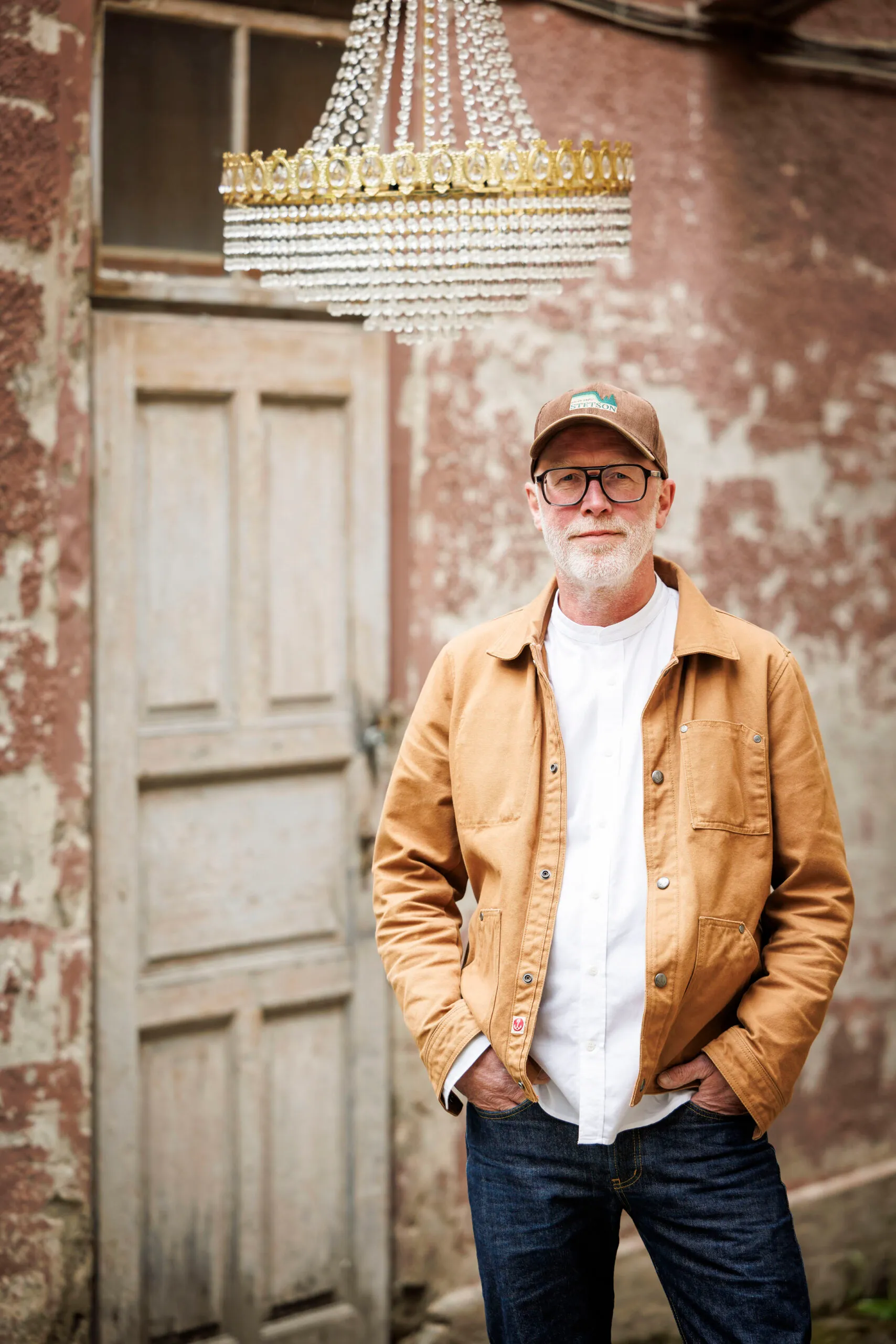 Mann mit brauner Jacke, weißem Hemd und brauner Cap steht mit den Händen in der Hosentasche in einem heruntergekommenen Hausflur. Hinter ihm eine alte helle Tür zu sehen, er schaut ernst in die Kamera