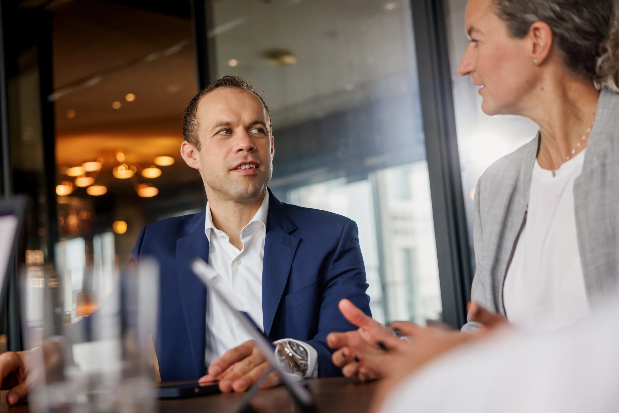 Zwei Personen sitzen an einem Konferenztisch, links ein Mann in einem blauen Sakko mit weißem Hemd und rechts im Anschnitte eine Frau mit grauem Jacket, weißem Shirt, Tablet und Gläser unscharf auf dem Tisch, im Hintergrund unscharf Glasflächen und Deckenlicht zu sehen