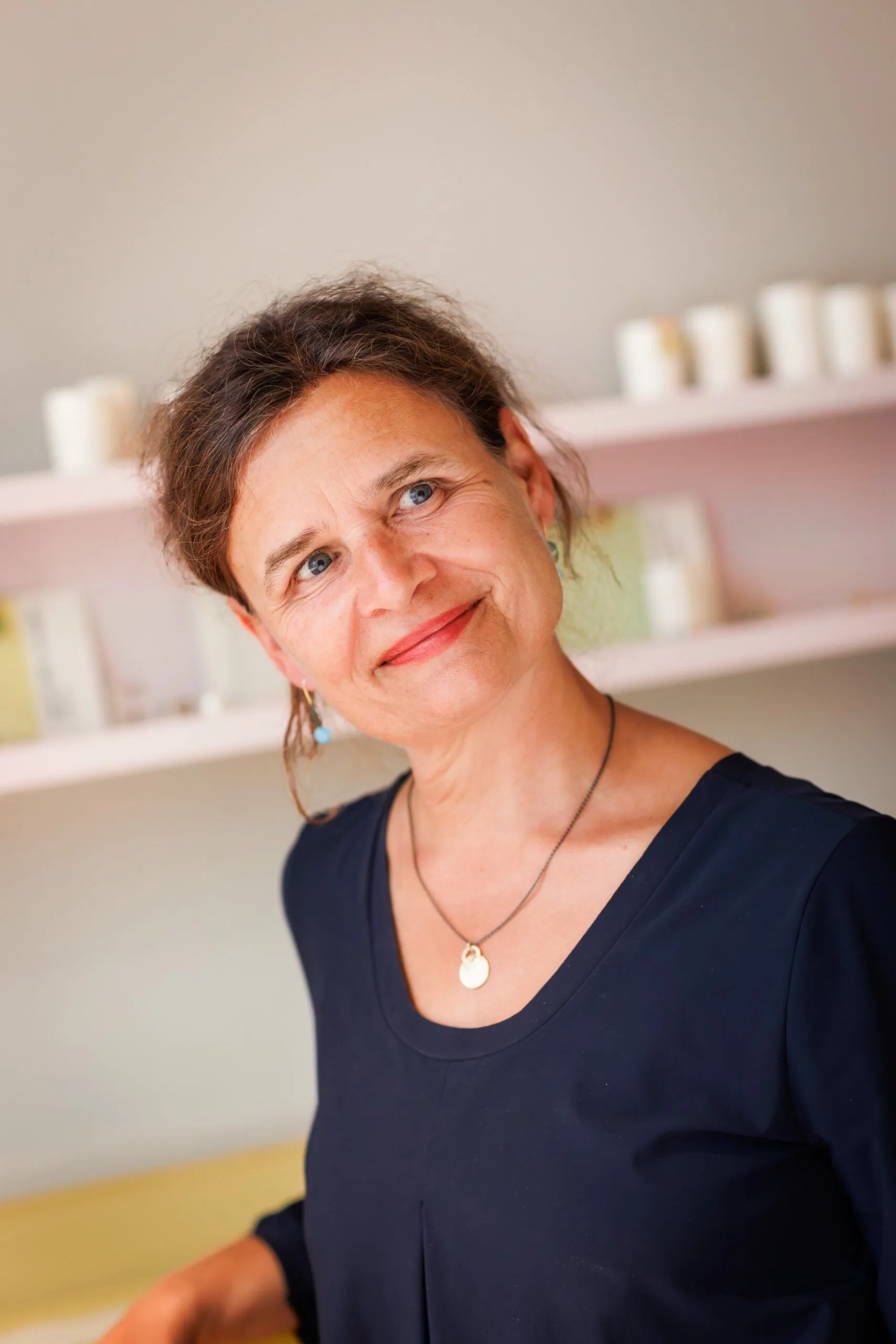 Portraits einer Goldschmiedin, sie trägt ein blaues Kleid, roten Lippenstift und schaut leicht zur Seite. Sie hat ihr braunes Haar in einem Zopf gebunden und trägt Ohrringe mit hellblauem Stein und eine Kette. Im Hintergrund unscharf eine helle Wand mit zwei Regalen.