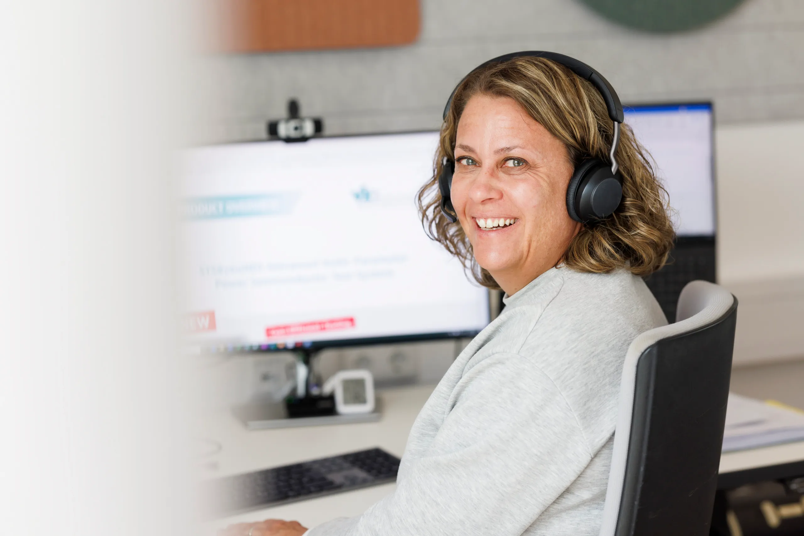 Eine Frau mit lockigem Haar und offenen Lachen trägt ein Headset und lacht in die Kamera. Sie sitzt von einem Bildschirm weggedreht.