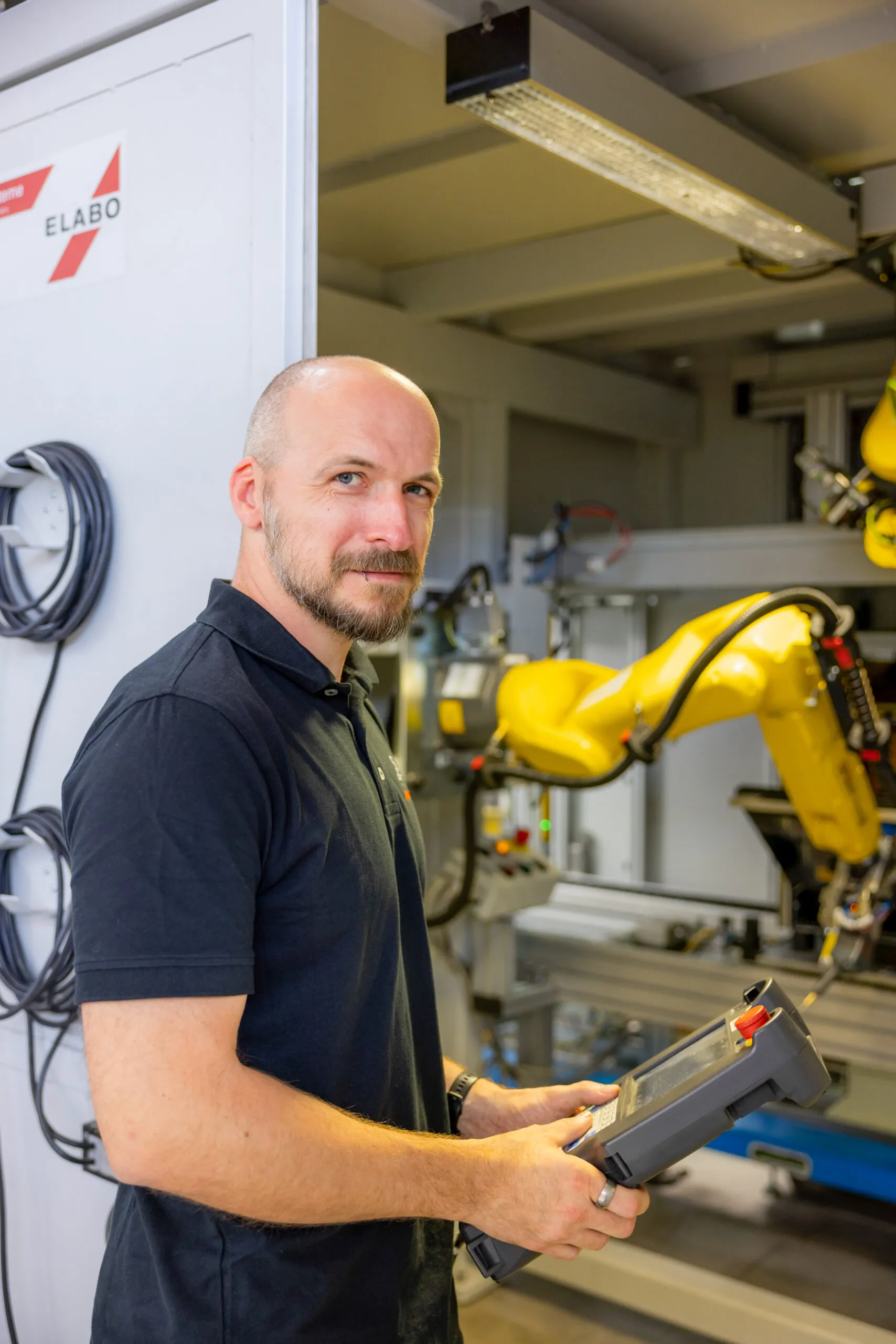 Ein Mitarbeiter mittleren Alters mit Halbglatze, Vollbart und einem blauen Poloshirt ist mit dem Oberkörper abgebildet. Er steht leicht nach rechts gedreht, mit einem leichten Lächeln und hat eine große Fernbedienung in der Hand. Im Hintergrund blickt man auf einen gelben Roboterarm. Links hängen aufgerollte Kabel an einer grauen Wand.