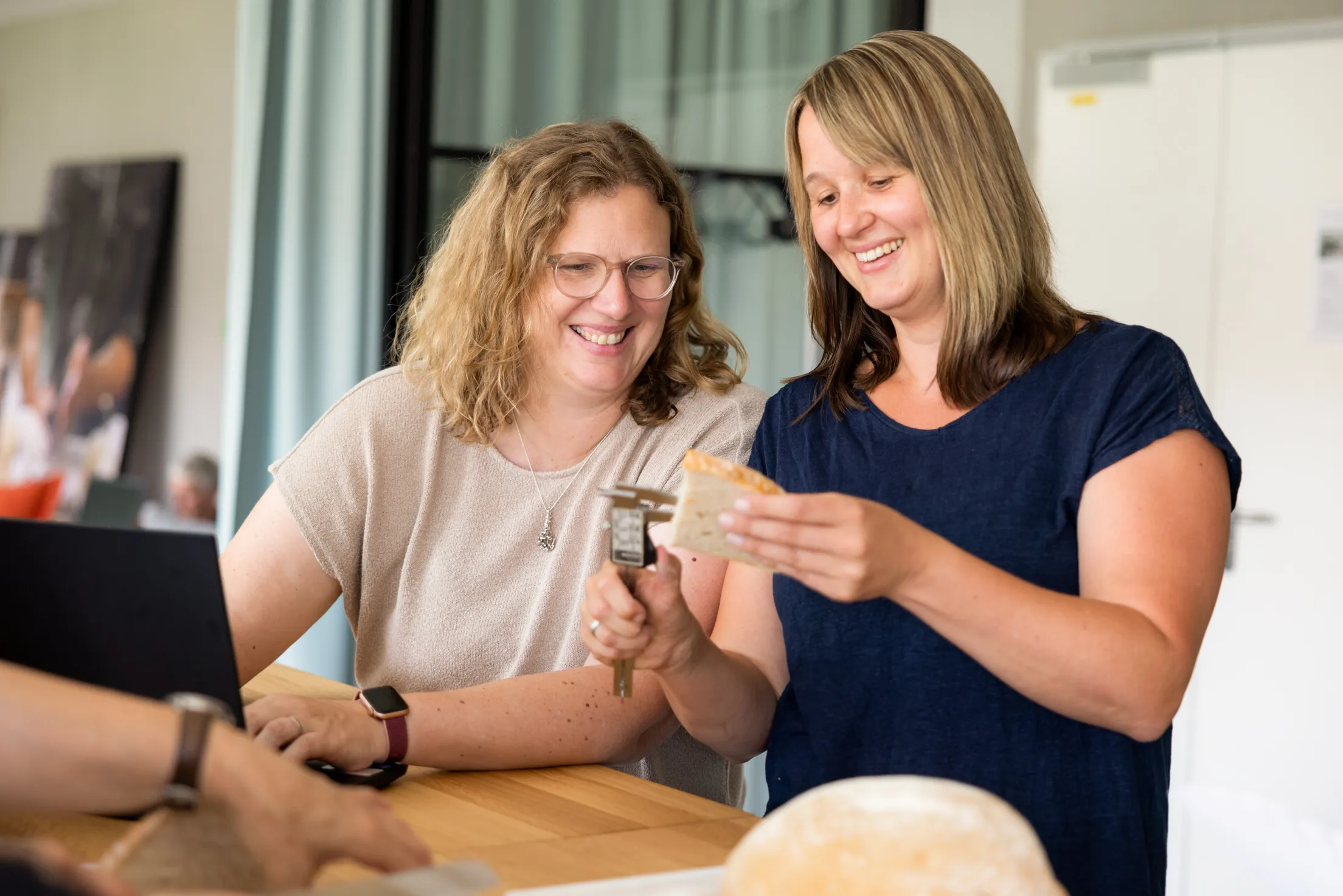 Zwei jüngere Damen stehen an einem Holztisch und testen ein Stück Brot. Die Dame links im Bild trägt eine helle Brille, hat mittelblonde, lockige, längere Haare und ein beiges Shirt an. Sie hat beide Hände an einem schwarzen Laptop und schaut auf eine Scheibe Brot, welche von der anderen Dame gehalten wird, neben einem Maßschieber. Sie hat dunkelblonde, glatte, mittellange Haare und trägt ein dunkelblaues Shirt. Im Hintergrund unscharf ein heller Raum zu erkennen.