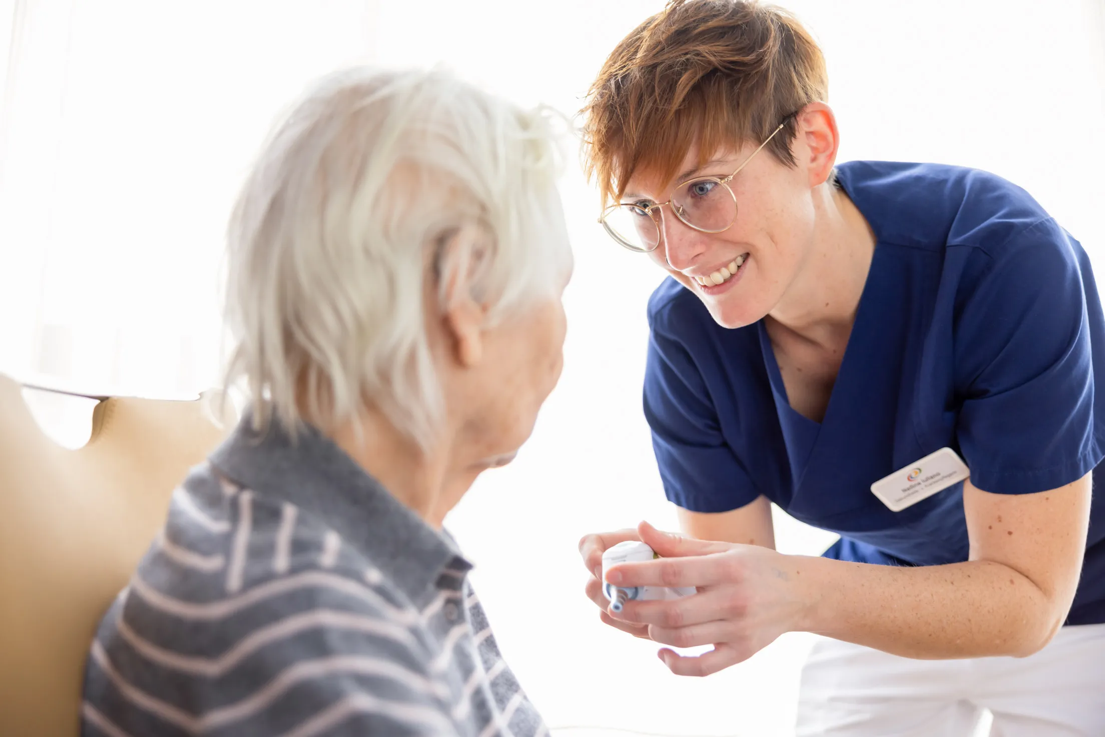 Das Foto ist ein Close-Up auf zwei Damen: links im Bild ist unscharf eine ältere Patientin mit weißen, kurzen Haaren, von der Seite fotografiert. Sie trägt einen gestreiften, grauen Pullover und schaut eine Pflegerin an. Diese steht rechts im Bild und beugt sich leicht herunter zu der Patientin. Sie trägt kurzes, kupferrotes Haar, eine goldene Brille und einen dunkelblauen Kasack. Sie lächelt und hat ein Fiebermessgerät in den Händen.