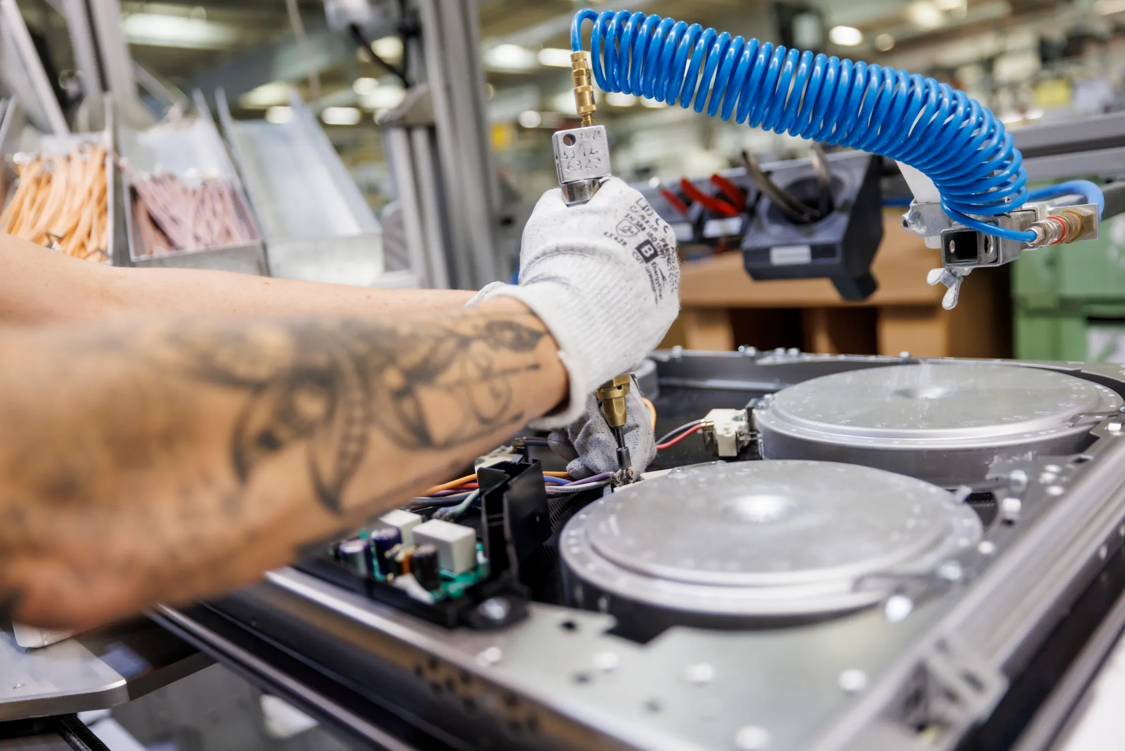 Detailaufnahme von einer Herdplatte an einem Fließband. Eine tätowierte Hand mit weißem Handschuh schraubt an der Herdplatte.