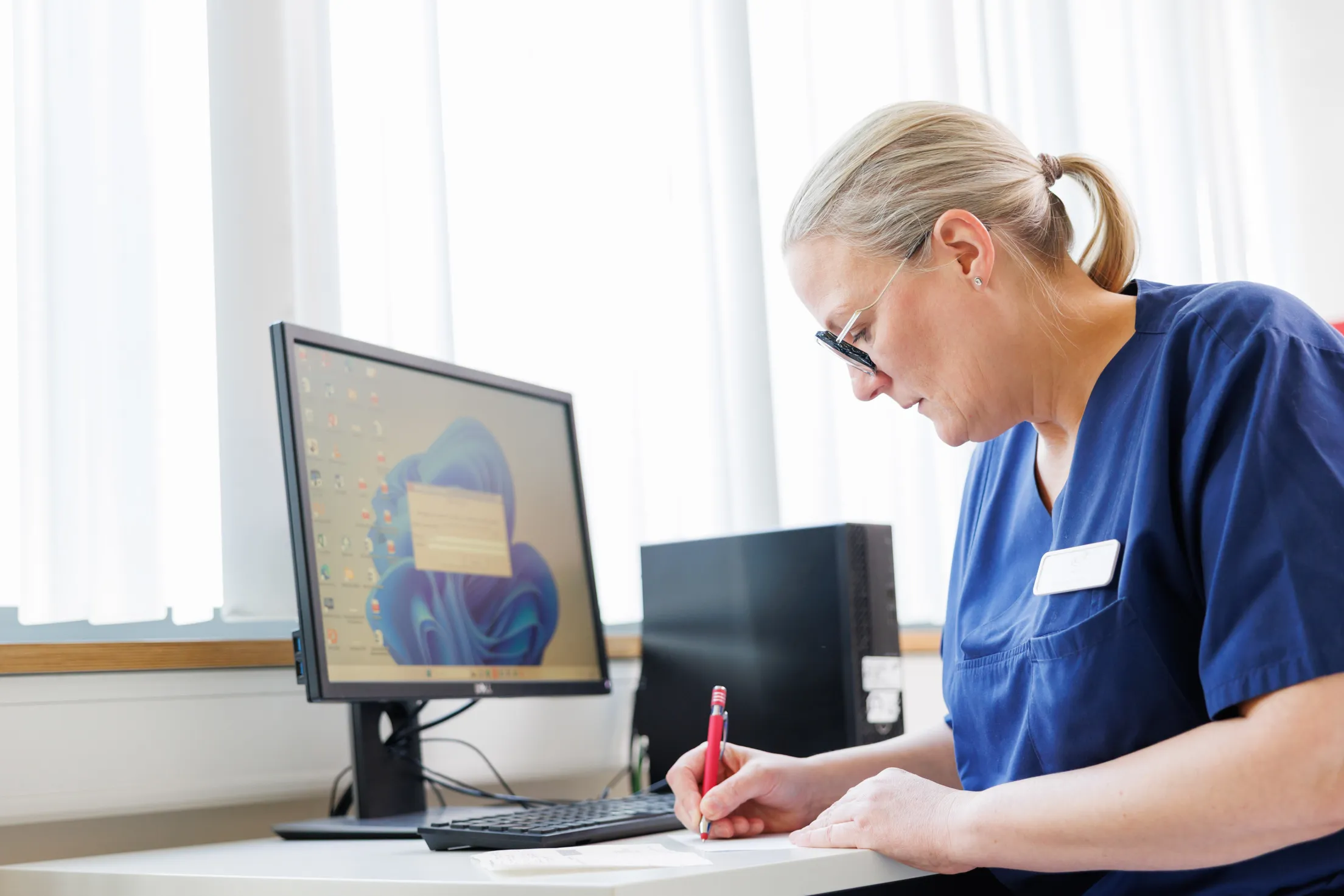 Blick auf einen weißen Schreibtisch mit einem schwarzen Computer, Monitor und Tastatur. Rechts im Bild eine Ärztin von der Seite mit ihrem Oberkörper zu sehen. Sie trägt einen blauen Kasack, blonden Pferdeschwanz und eine auffällige schwarze Brille. Sie schreibt etwas auf ein Blatt Papier, in der rechten Hand einen roten Kugelschreiber.