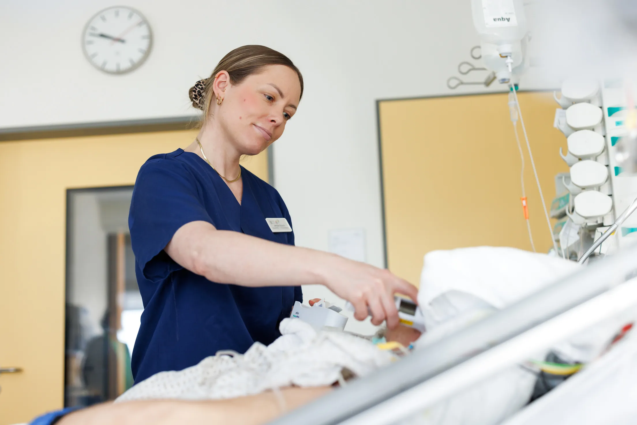 Eine Krankenpflegerin mit dunkelblondem Haar, streng zu einem Dutt zusammengebunden steht am Bett eines Patienten auf der Intensivstation. Sie trägt einen blauen Kasack und misst gerade Fieber am Patienten. Der Patient ist nicht zu sehen, man sieht nur das Patientenbett im Anschnitt, das Kopfende nach oben gefahren und rechts mit Infusionsflaschen, Schläuchen und Überwachungsgeräte. Im Hintergrund sind zwei gelbbeige Türen und eine Wanduhr zu sehen.