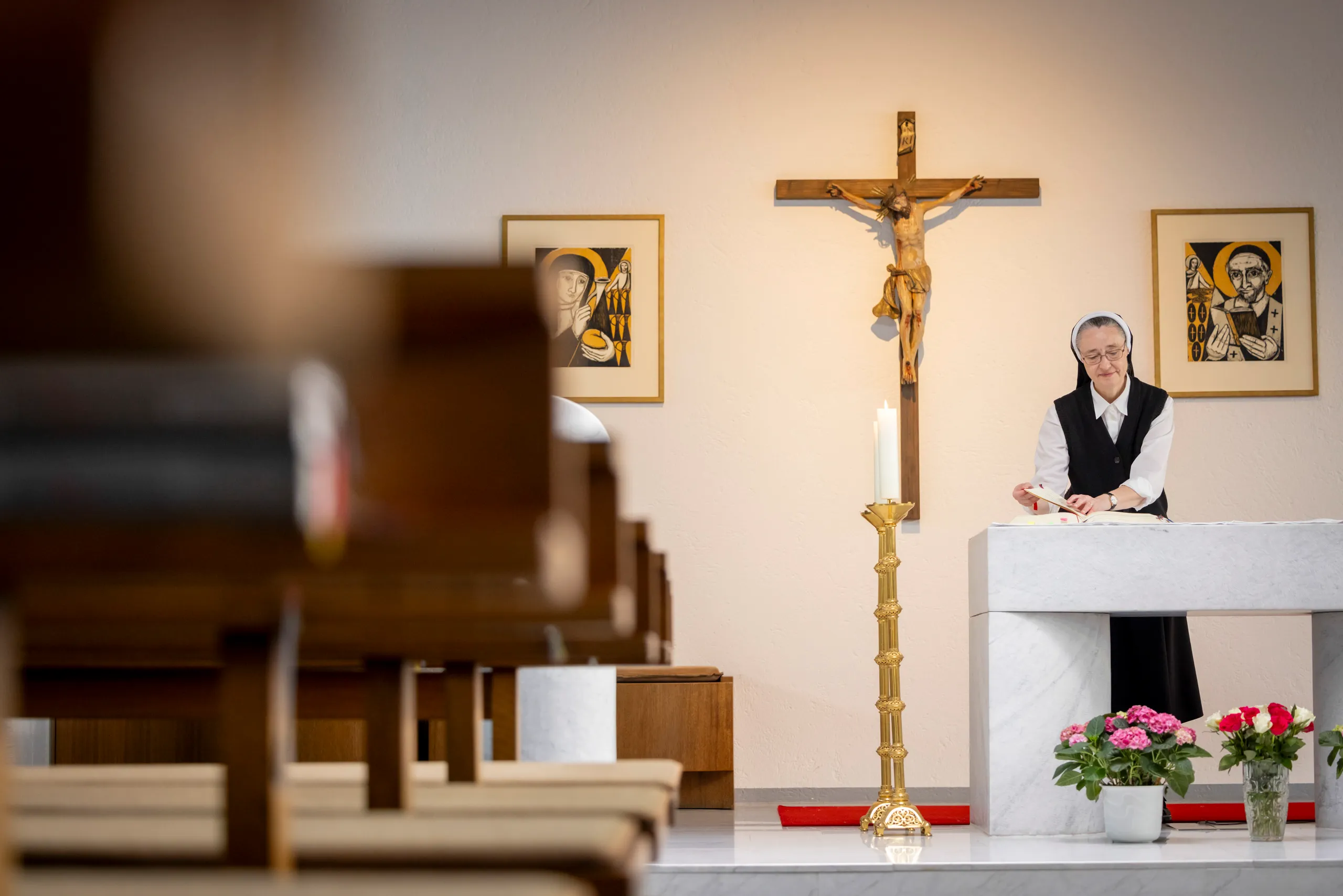 Eine Ordensschwester ist rechts im Bild an einem Altar zu sehen. In der Mitte ein großes Kreuz und unscharf sind Kirchenbänke zu sehen.