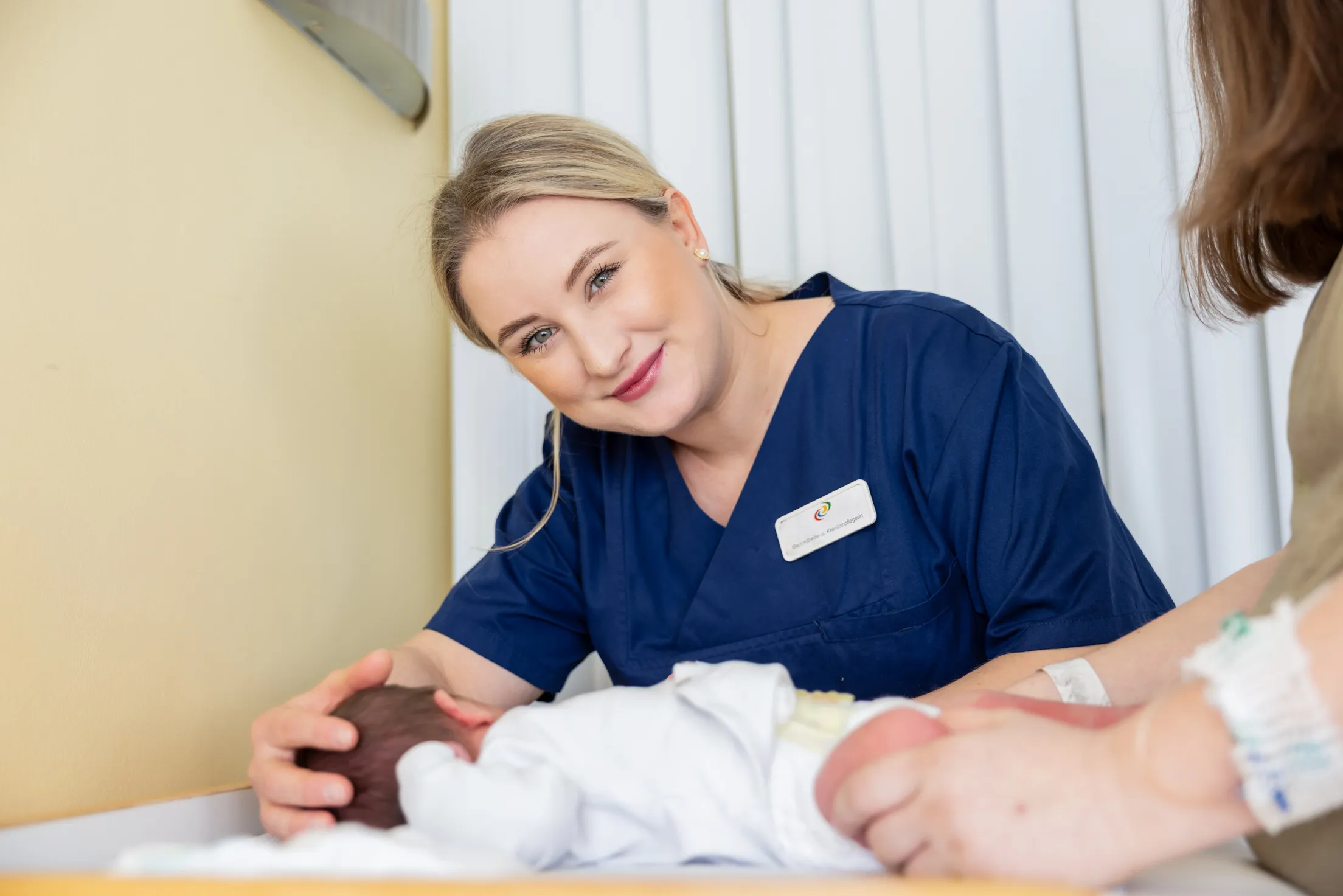 Eine jüngere Krankenpflegerin mit blonden Haaren zu einem Pferdeschwanz gebunden, trägt roten Lippenstift, einen dunkelblauen Kasack und lächelt in die Kamera. Sie beugt sich über ein Baby und hat ihre rechte Hand an dem Köpfchen des Kindes. Das Kind ist in einem weiße Body und die Mutter ist rechst im Bild nur im Anschnitt zu sehen. Sie trägt ein olivgrünes Shirt und hat braune, kinnlange Haare.
