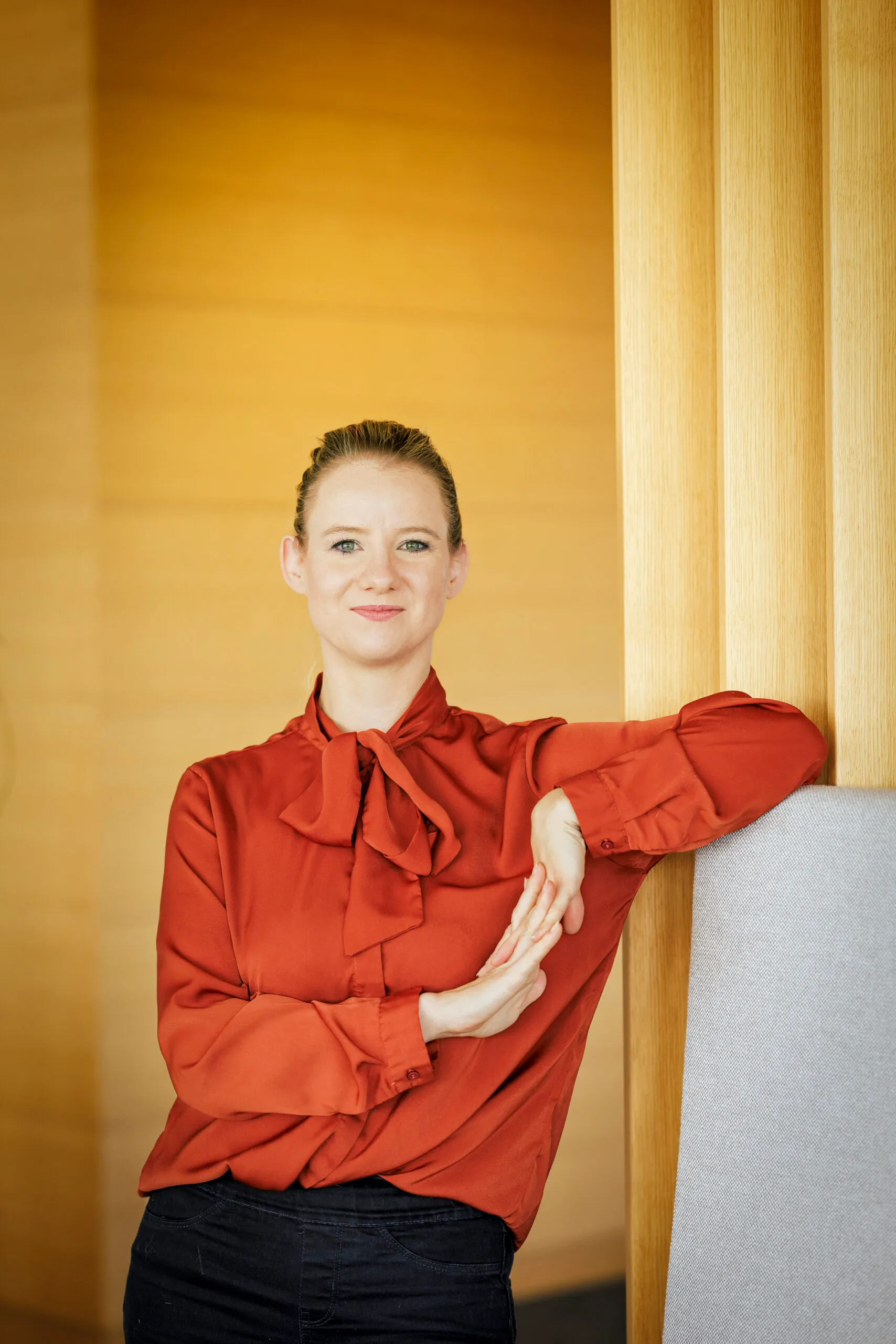 Portraits einer jungen Dame, die ihre dunkelblonden Haare zu einem strengen Zopf trägt. Sie trägt eine knallige, orangefarbene Bluse und macht eine schöne Wellenbewegung mit ihren Händen angelehnt an eine graue Lehne