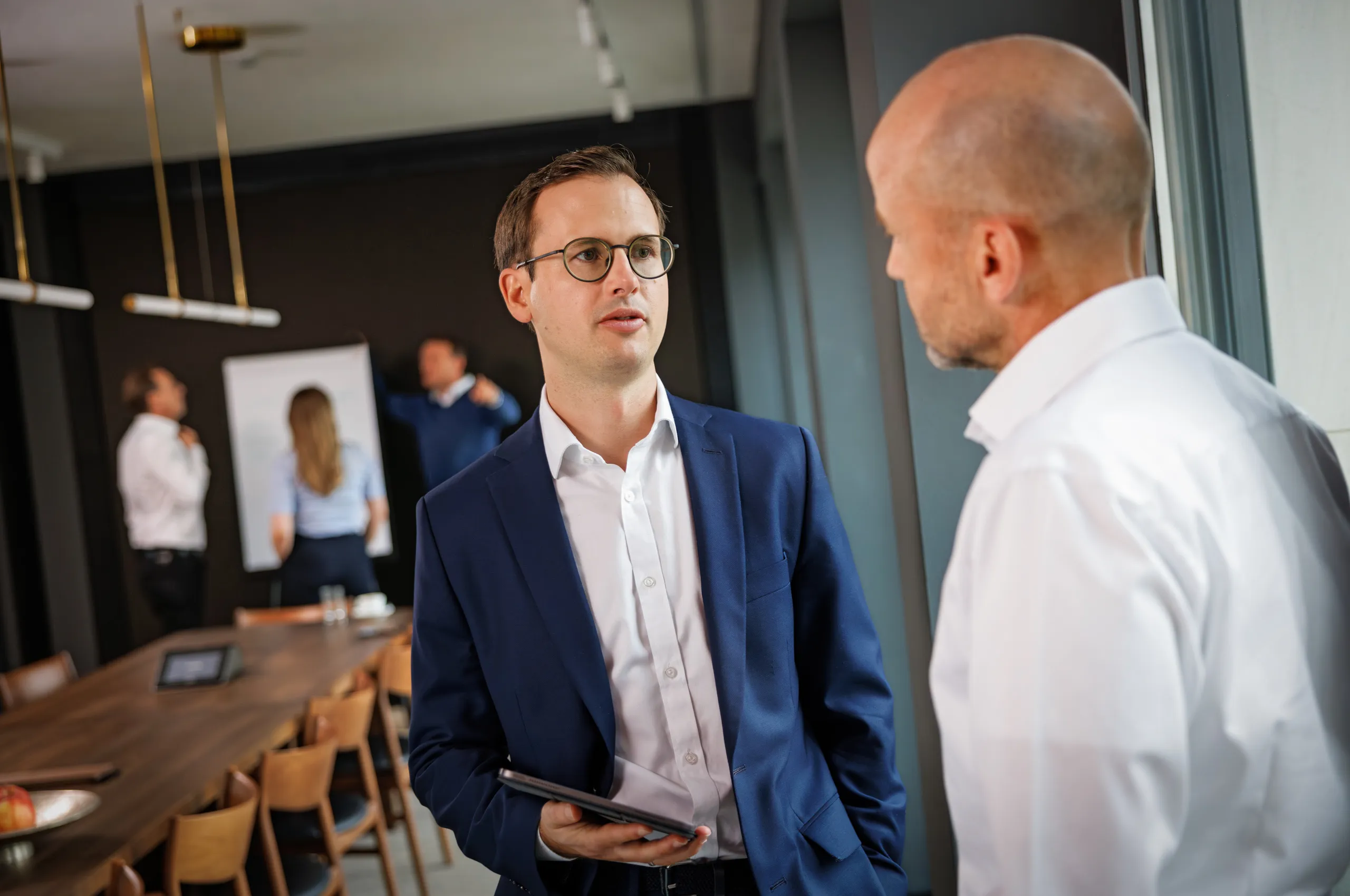 Zwei Business Männer rechts im Bild sind im Gespräch. Links ein Herr im dunkelblauen Anzug, weiße, Hemd und mit einem Tablet in der Hand, rechts ein Mann mit kahlem Kopf unscharf in einem weißen Hemd. Im Hintergrund unscharf ist ein langer Holztisch zu sehen und am Ende des Tisches drei Personen an einem Flipchart. Die Wand ist schwarz.