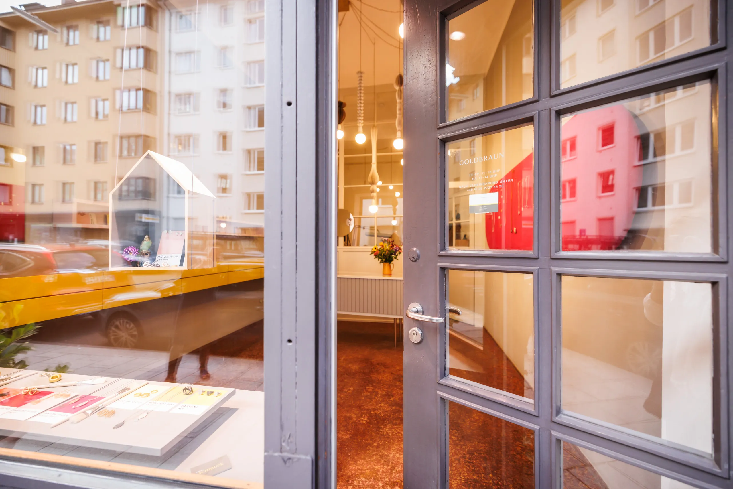 Eine graue Eingangstür aus Glas ist von der Strasse aus zu sehen, die leicht geöffnet ist und den Blick in den Schmuckladen führt. Links unscharf ein Schaufenster mit Schmuck, Spiegelung im Fenster. Ein rotbrauner Fussboden ist zu sehen und auf der Tür steht Goldbraun und die Öffnungszeiten.