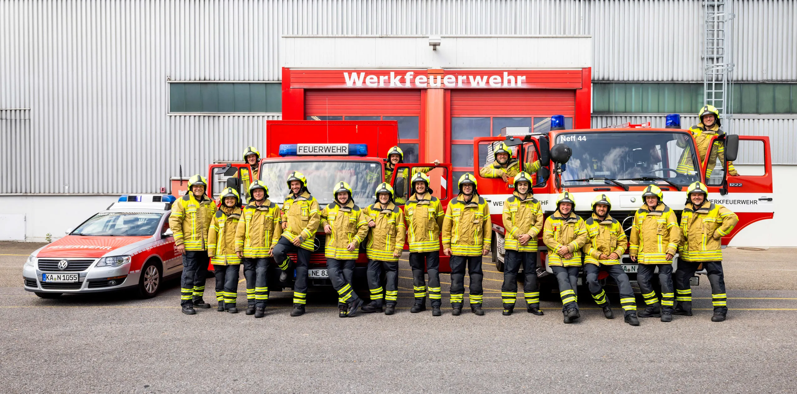 Ein Gruppenfoto der Werksfeuerwehr draussen vor dem roten Eingangstor des Gebäudes mit zwei roten Feuerwehrwägen und einem Einsatzfahrzeug der Feuerwehr. Die Mitarbeiter alle in voller Montur mit gelben Jacken und Helmen, schwarzen Hosen, stehen in einer Reihe vor den Wägen, teilweise auch seitlich in den Autos bei Fahrer- und Beifahrertür. Alle schauen in die Kamera.