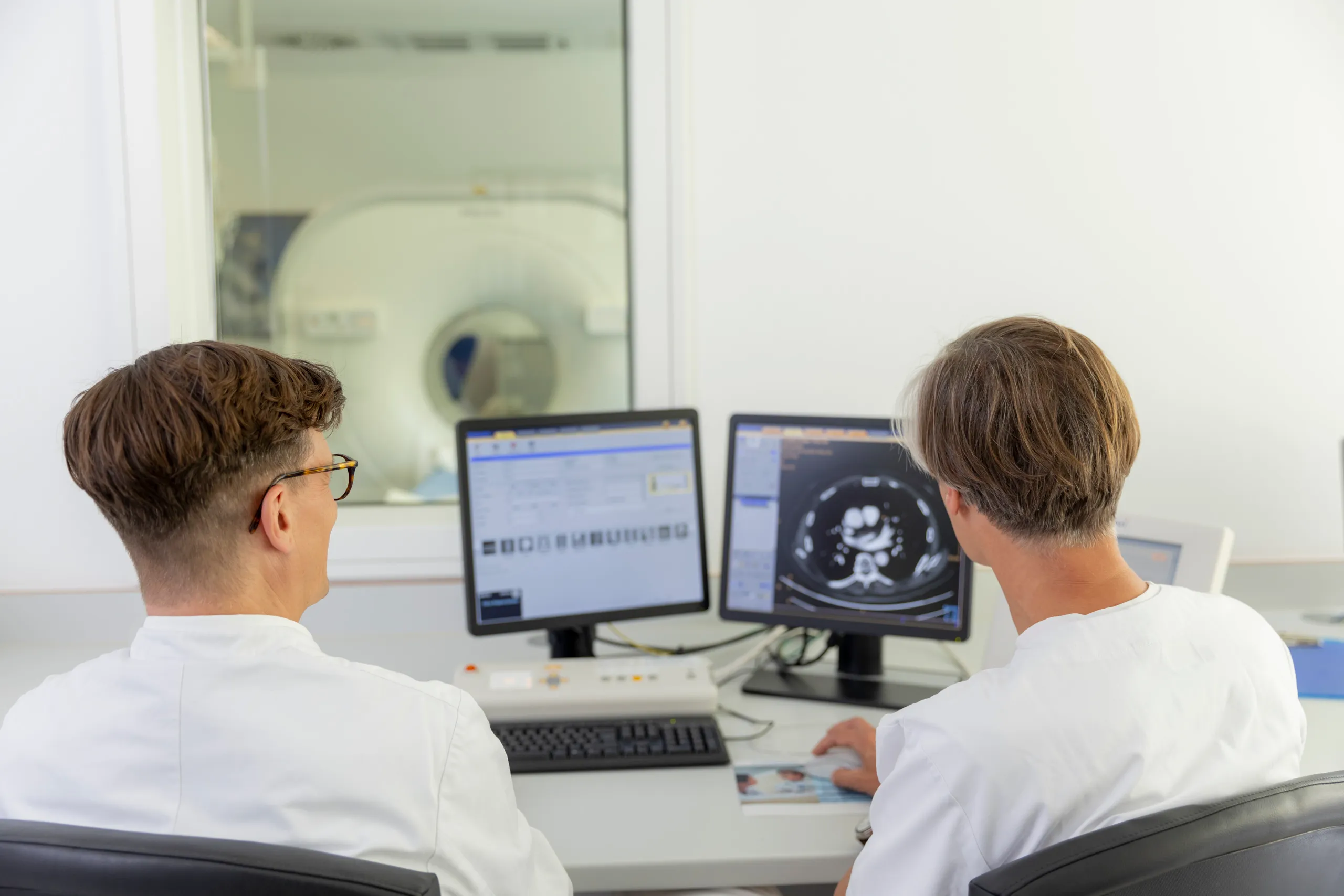 Zwei Ärzte sitzen mit dem Rücken zur Kamera und betrachten Röntgenbilder auf zwei Monitoren. Die Computer und im Hintergrund ein Kernspingerät sind unscharf zu sehen.