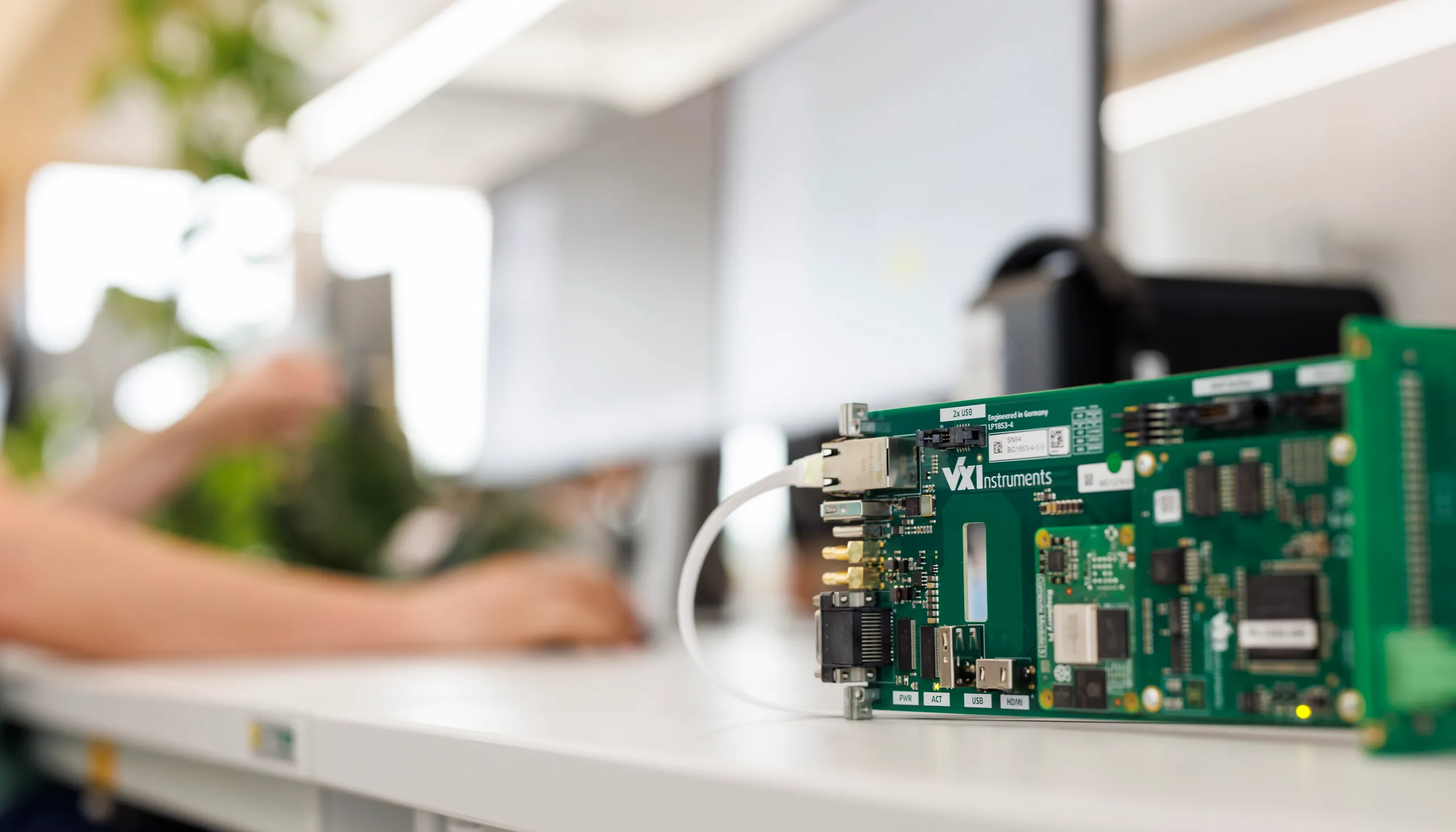 Auf einem weißen Schreibtisch steht ein Halbleitersystem ohne Verdeckung. Im Hintergrund links im Bild sieht man unscharf zwei Arme von einem Mitarbeiter.