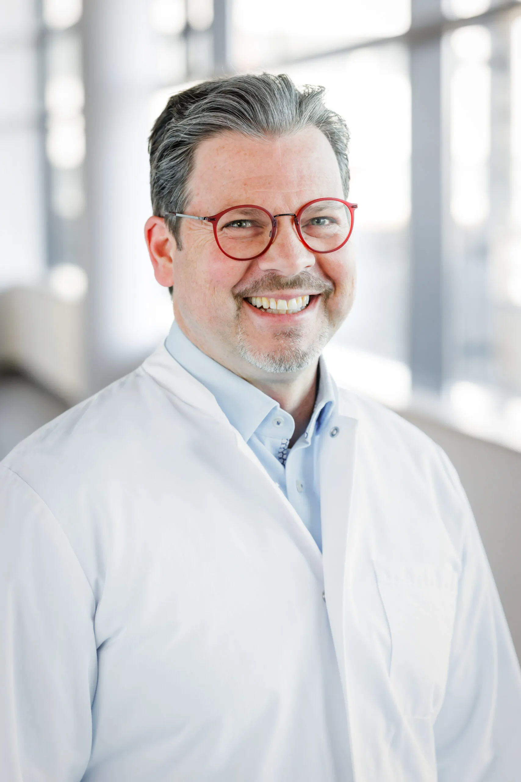 Hochformatiges Portrait eines Arztes mittleren Alters. Er trägt ein hellblaues Hemd und darüber einen weißen Kittel. Er hat graumeliertes kurzes, welliges Haar, Bart und eine rote, runde Brille. Er lacht in die Kamera und ist mit dem Oberkörper abgelichtet. Im Hintergrund ist unscharf ein heller Klinikgang mit Säulen und Fenster zu erkennen.