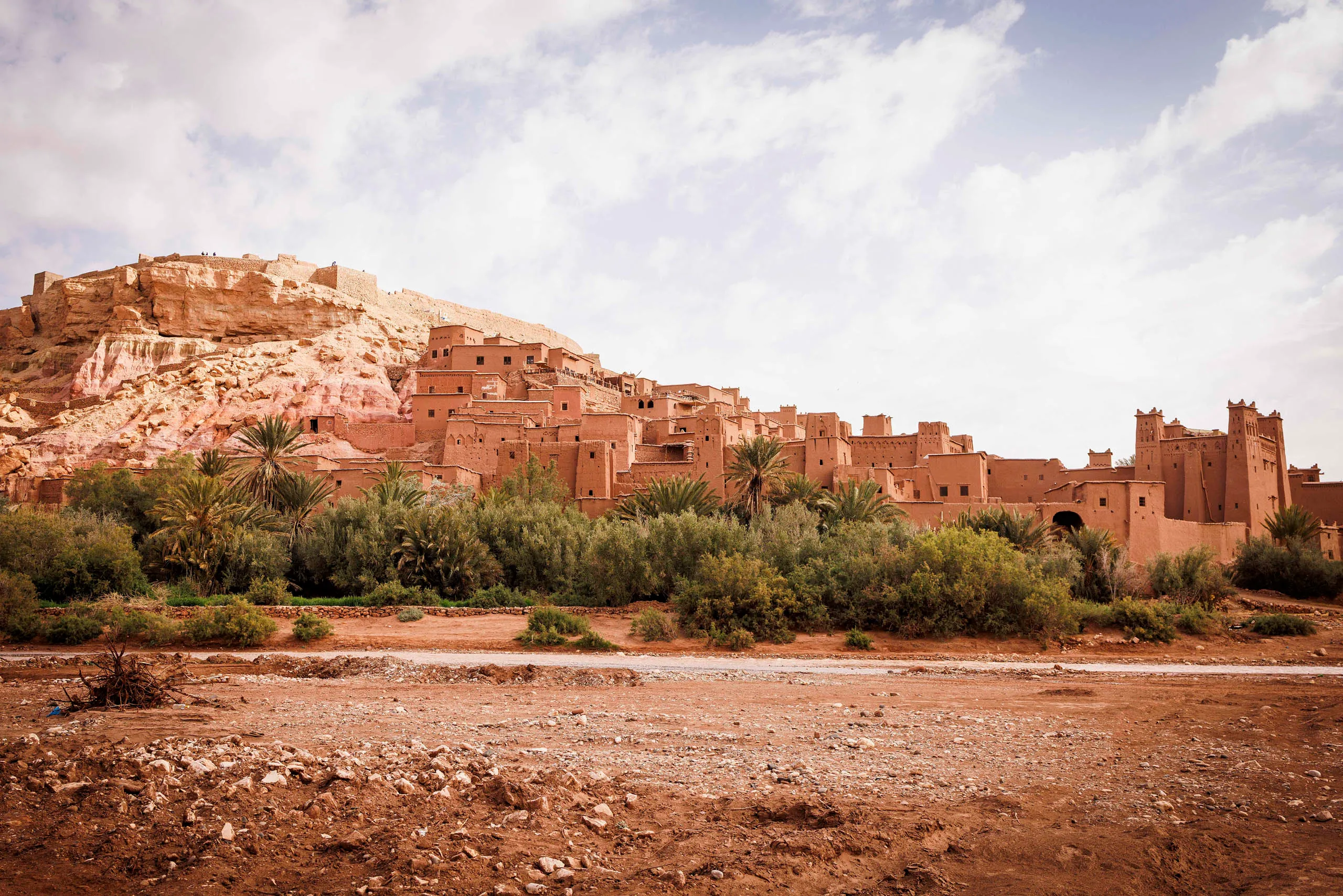 Die Lehmstadt Aît Ben Haddou mit dem Aussichtshügel links und Palmen ist abgebildet. Davor fliesst ein kleiner Fluss und Wüstenboden grenzt vorne im Bild an.