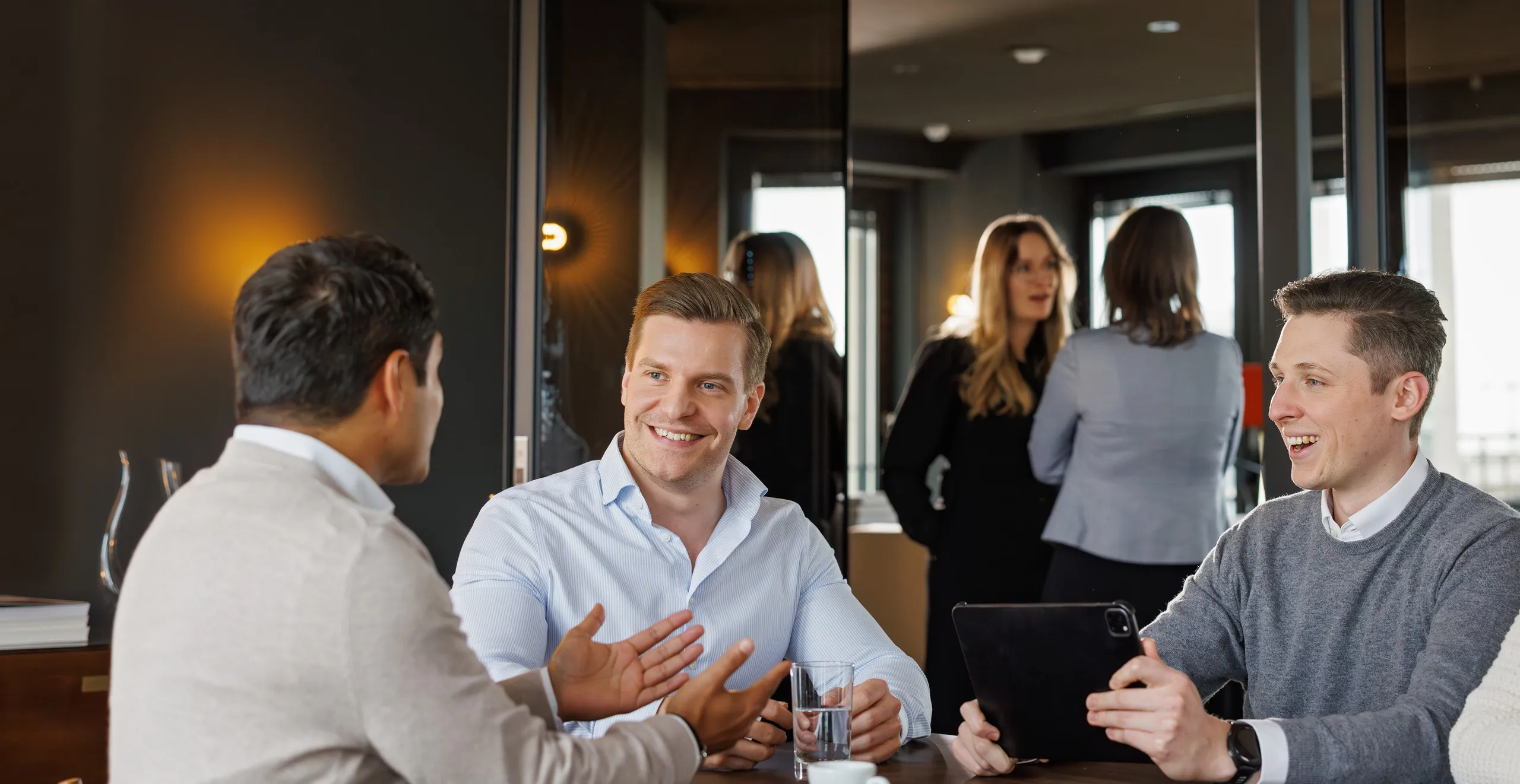 Drei jüngere Männer sitzen an einem Tisch bei einer Besprechung mit Tablet, sie diskutieren und lachen zusammen. Links der Mann im Anschnitt trägt einen beigen Pullover und ein weißes Hemd, der Mann in der Mitte ein hellblaues Hemd und er lacht zu seinem Kollegen links im Bild. Rechts sitzt ein weiterer junger Mann mit weißem Hemd und grauem Strickpullover, er hält das Tablet fest. Alle drei haben kurze, dunkle Haare. Im Hintergrund stehen unscharf zwei Mitarbeiterinnen im Businessoutfit im Gespräch. Der Raum im Hintergrund ist sehr dunkel gehalten und hat große Fenster.