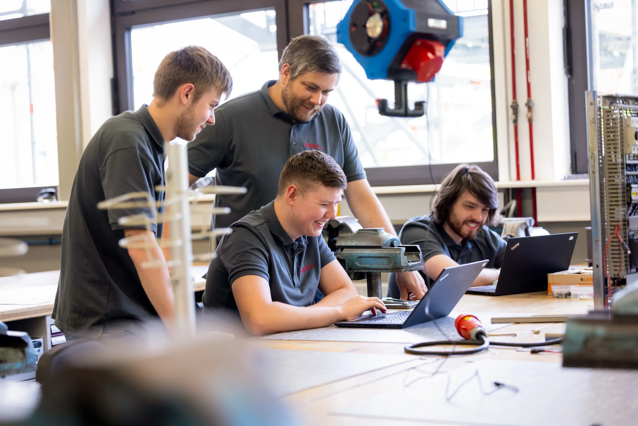In der Azubi Werkstatt mit vielen Fenstern sind drei Auszubildende zu sehen und ein Ausbilder mittleren Alters. Alle tragen ein dunkelgraues Neff Poloshirt mit rotem Neff Logo. Zwei der jungen Männer sitzen und schauen in zwei Laptops. Die anderen beiden stehen dahinter und schauen den beiden über die Schultern auch in den Laptop. Im Vordergrund unscharfe Elemente, zB eine große Steckdose, die an der Decke hängt.
